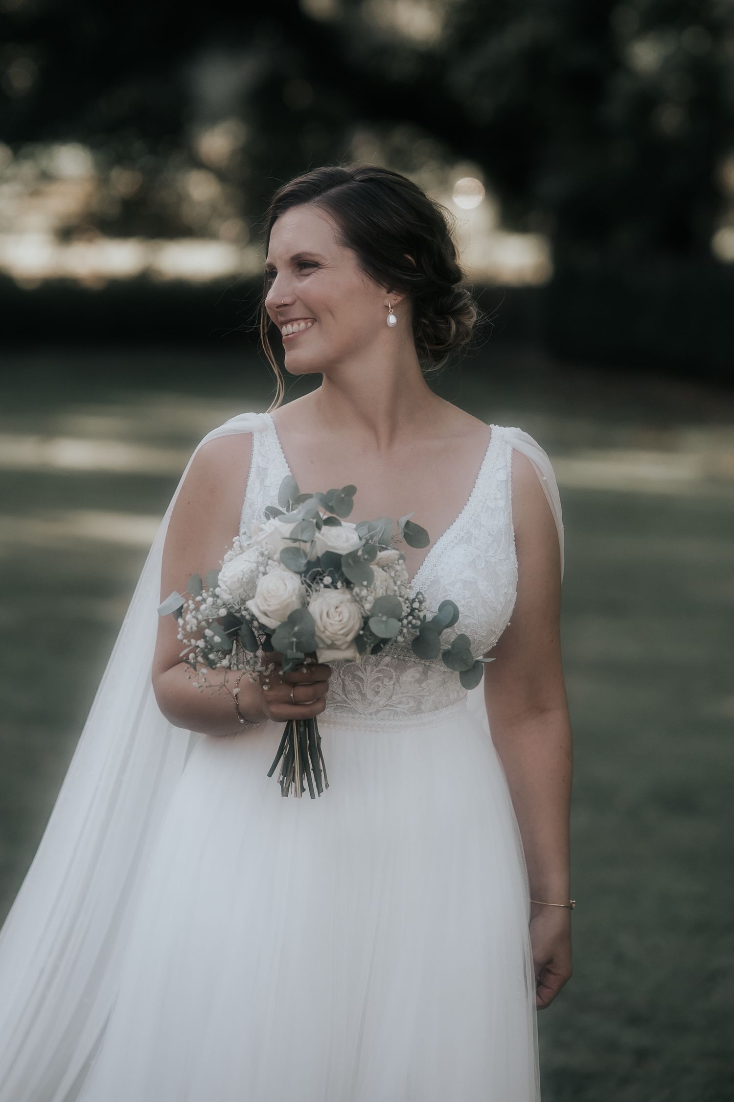 A bride in a flowing white dress holds a beautiful bouquet of white roses and eucalyptus against an outdoor backdrop.