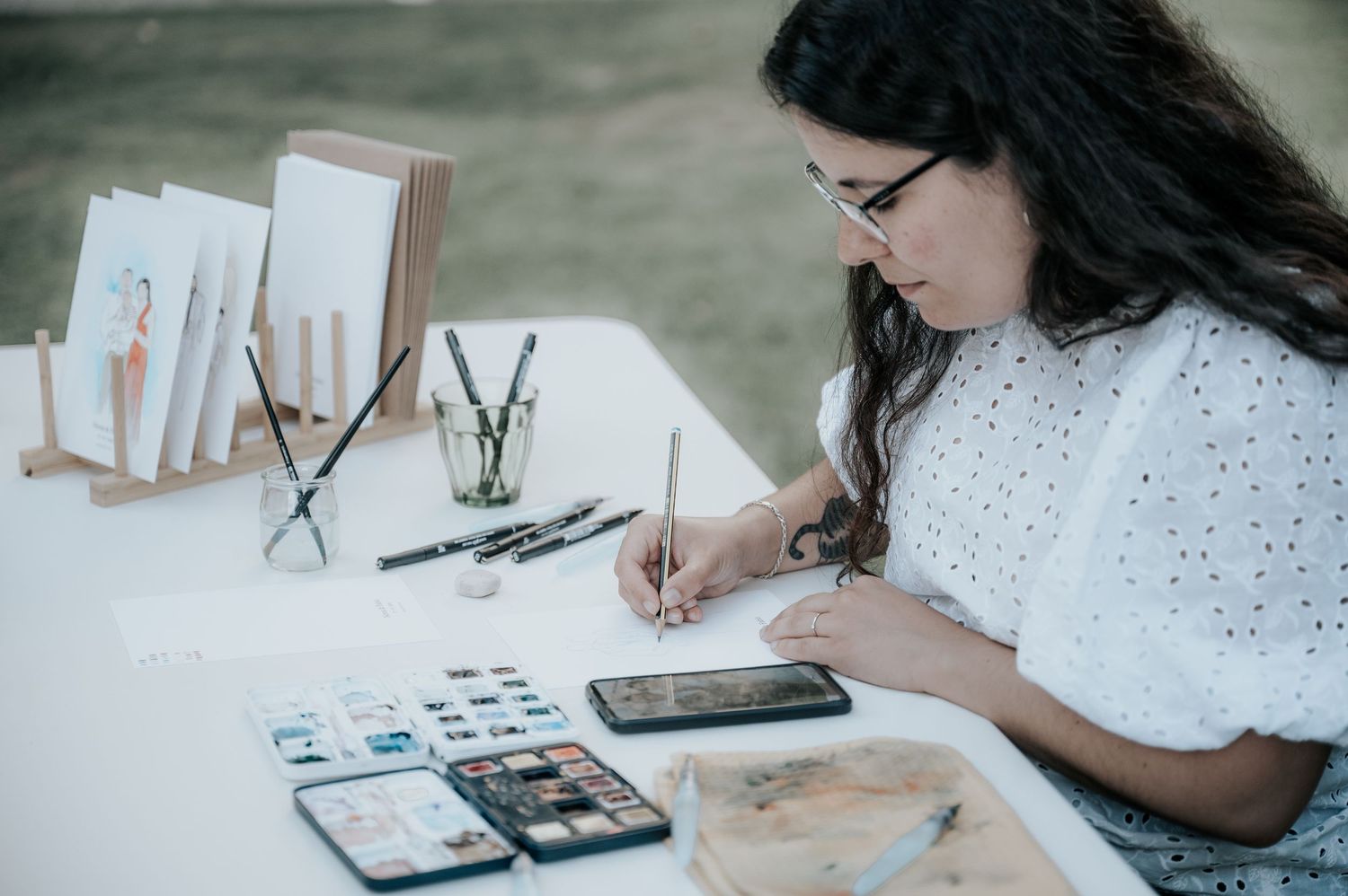 Artist working on watercolor sketches at desk with art supplies and reference materials spread out.