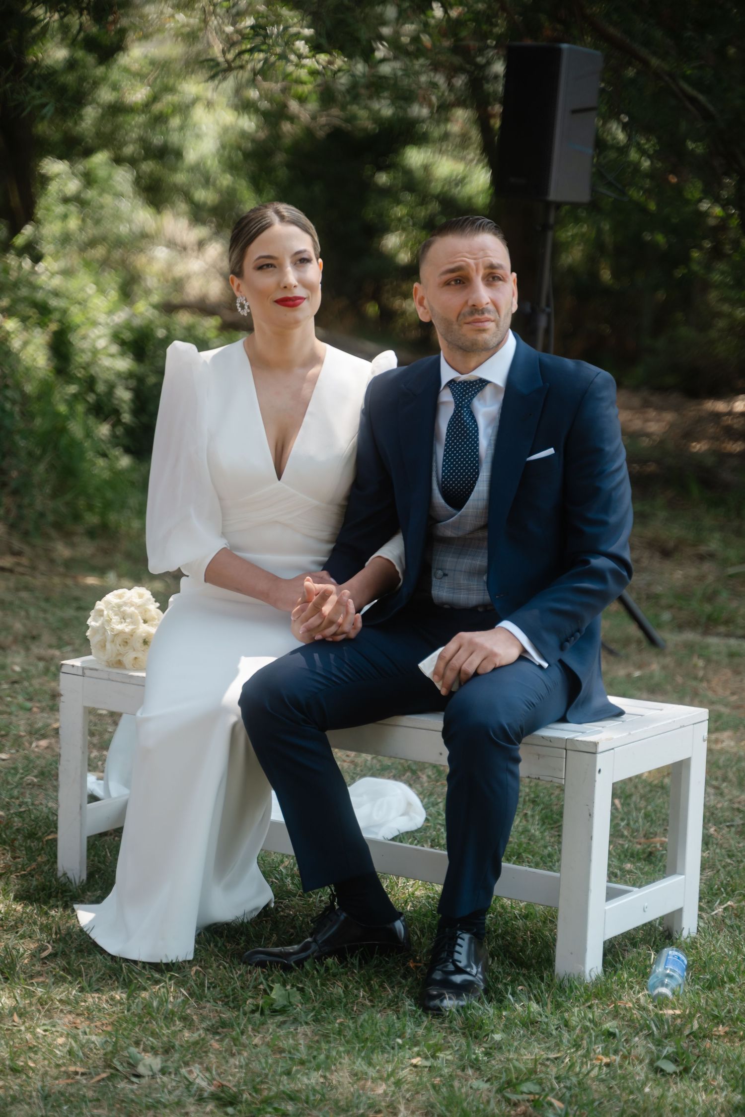 A couple in wedding attire sits together on white wooden benches in a garden setting.