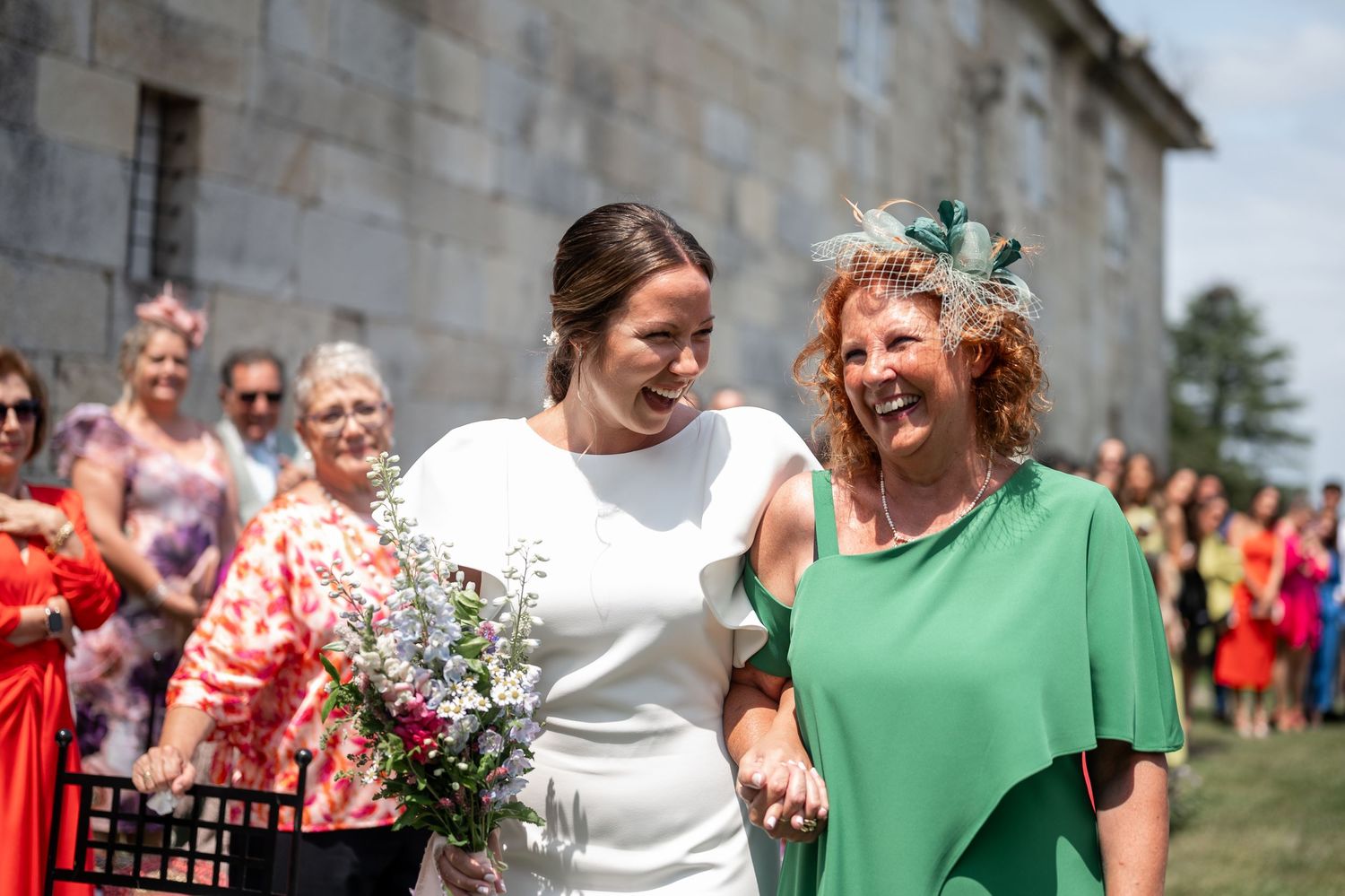 Two people in white and green dresses share a joyful moment at an outdoor celebration with guests in the background.