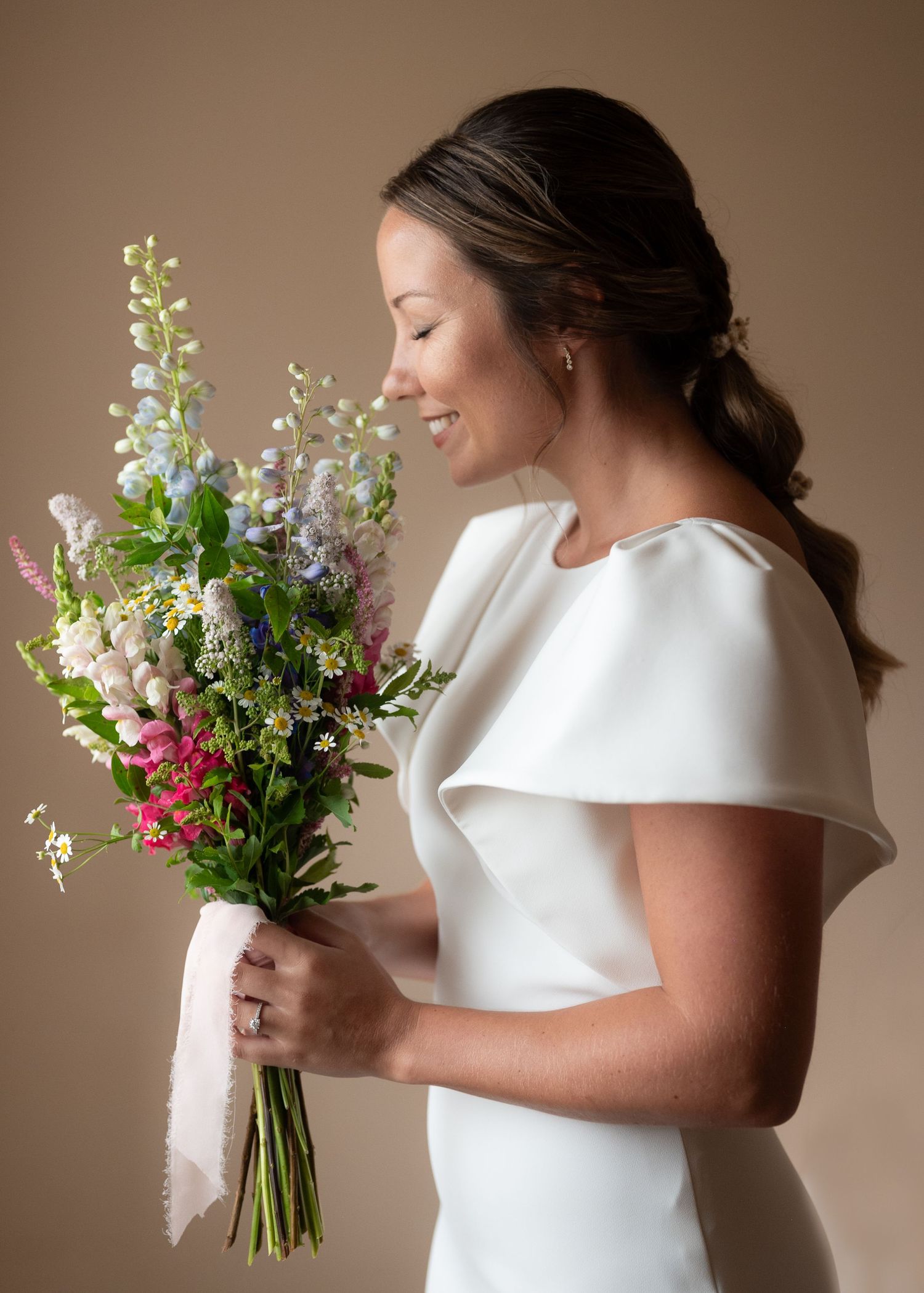 Woman in classic white dress admiring vibrant wildflower bouquet against beige background.