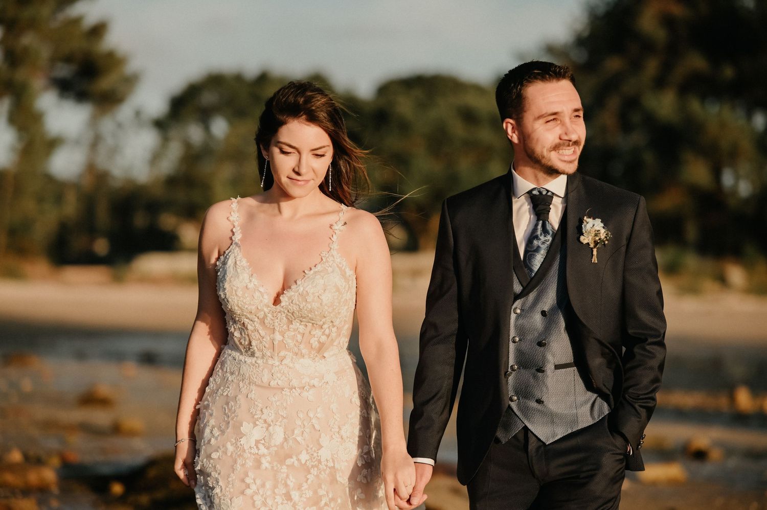 A newlywed couple walks hand in hand through a golden field during sunset, with the bride wearing a lace dress.