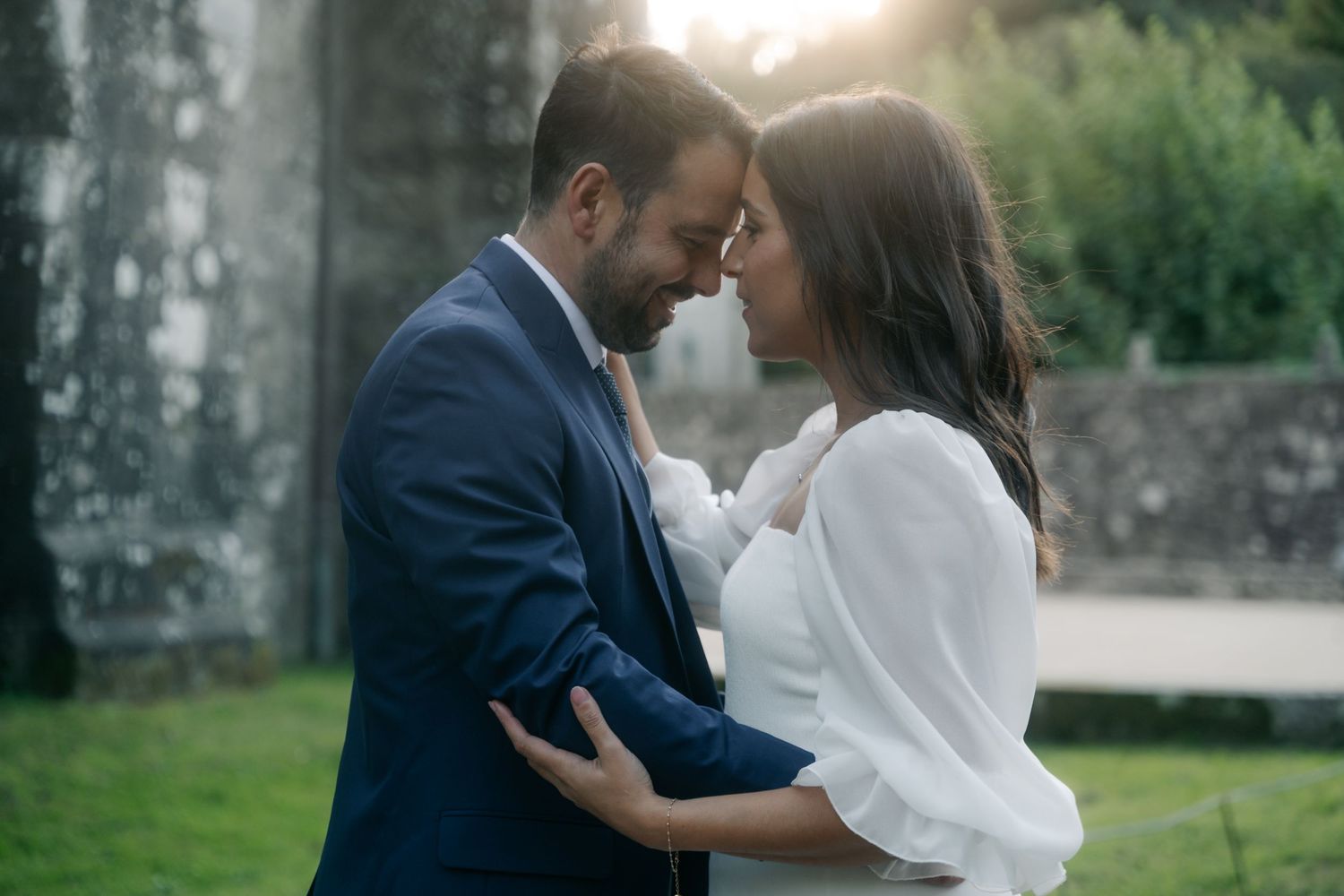 A couple in wedding attire shares an intimate moment outdoors at sunset in a garden setting.