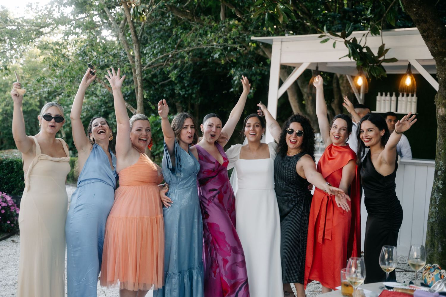 Wedding party group photo showing bridesmaids in various pastel and bright colored dresses standing together outdoors.