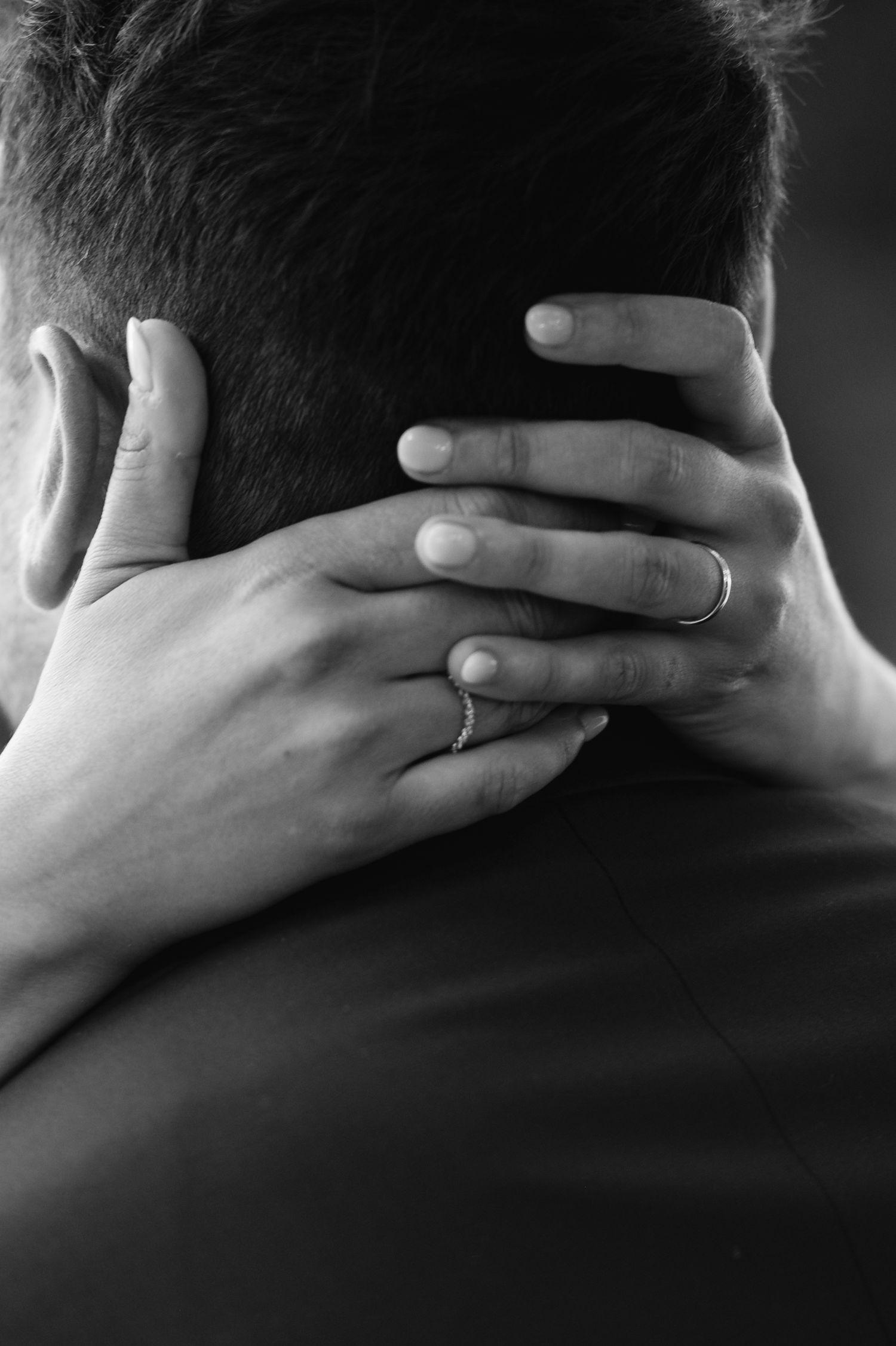 A black and white close-up of hands with a wedding ring embracing the back of a neck.