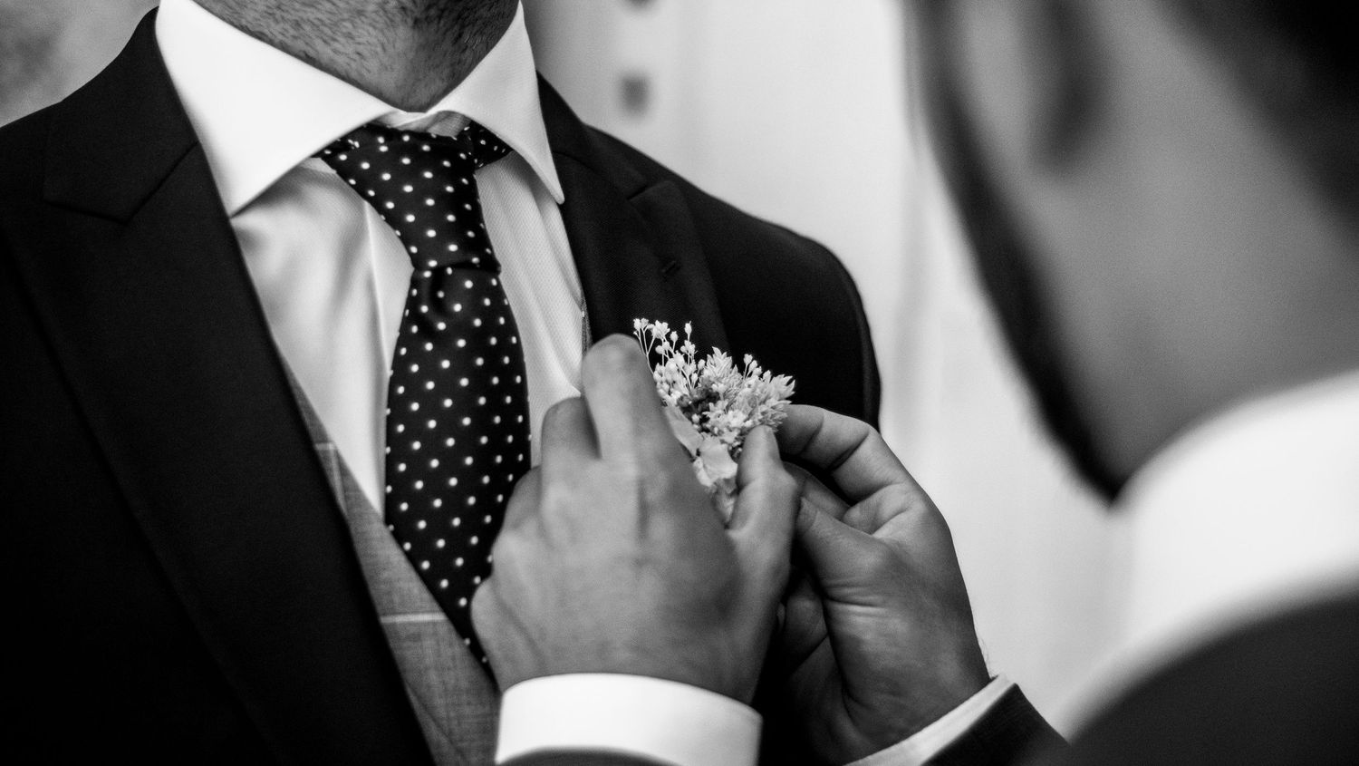 Black and white close-up of hands pinning a small floral boutonniere onto a polka dot tie and suit jacket.