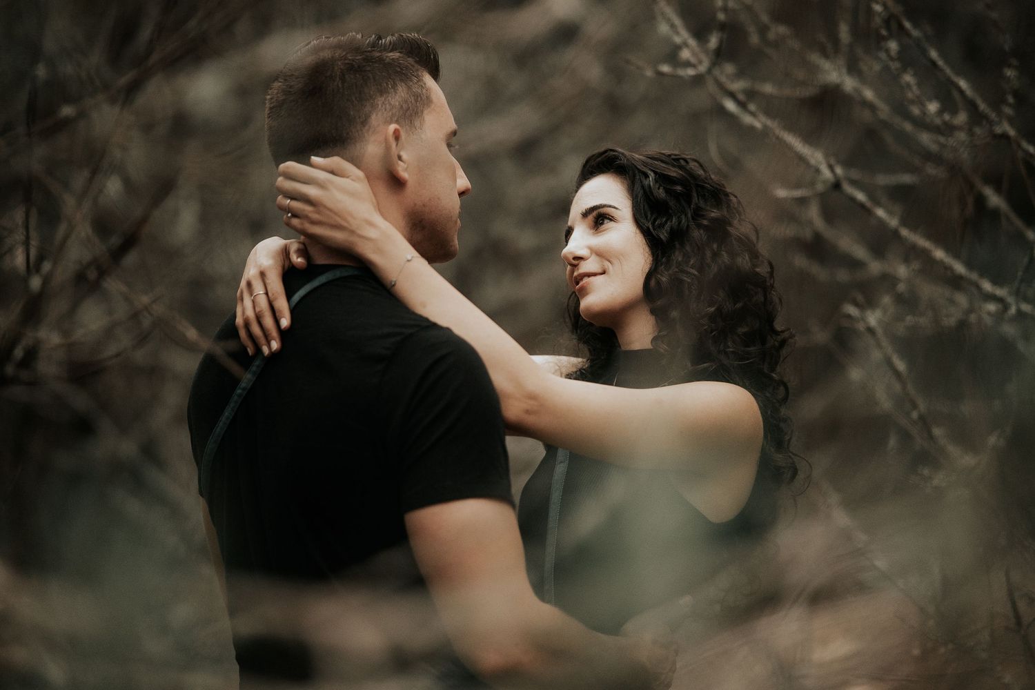 A couple embraces intimately in a moody forest setting with dramatic lighting and shadows.