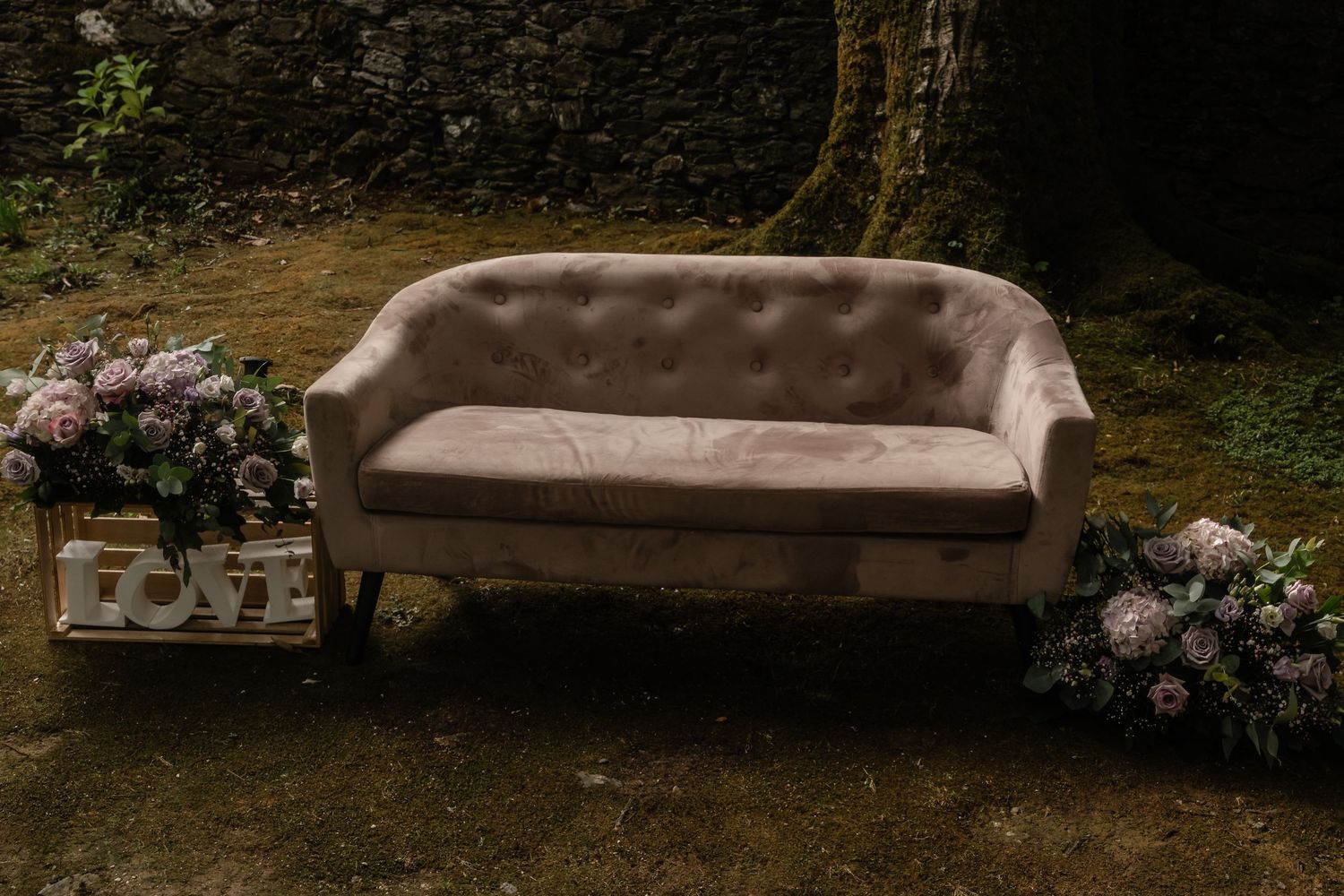 A beige couch sits outdoors in a garden setting surrounded by pink flowers and greenery.