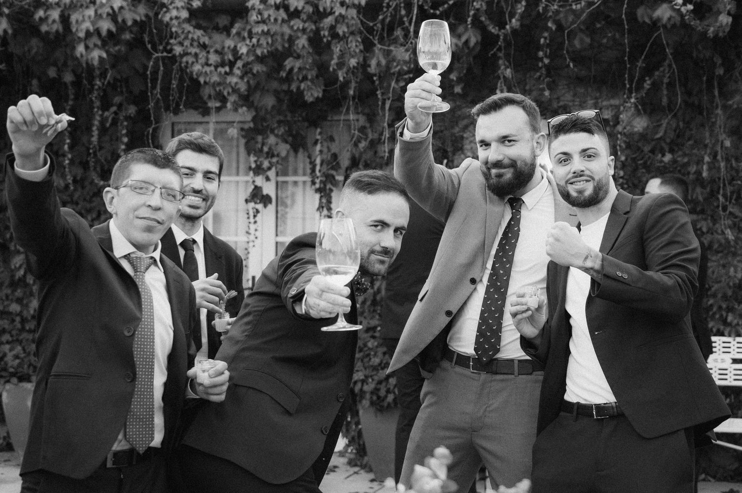 Group of groomsmen in dark suits toasting with drinks at an outdoor wedding celebration.