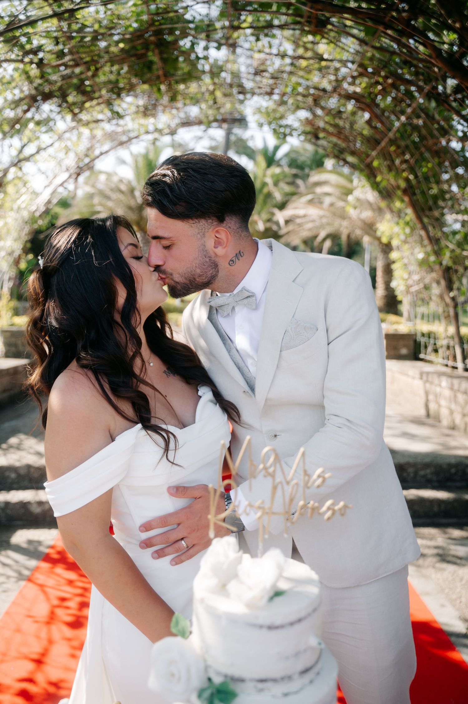 A couple in wedding attire shares romantic kisses while cutting their white wedding cake outdoors on a sunny day.