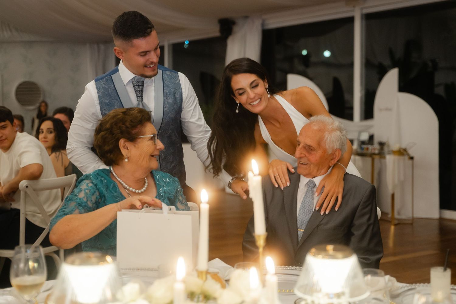 Wedding guests enjoy a candlelit dinner reception with laughter and conversation at an elegant indoor venue.