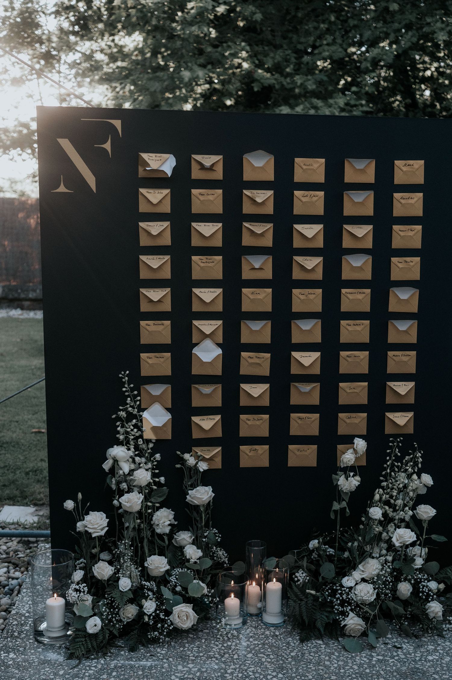 Wedding seating chart display with gold envelopes arranged on dark boards, decorated with white roses and candles.