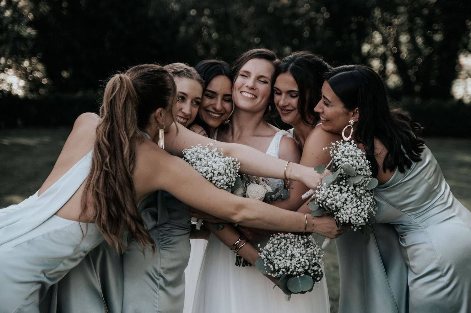 Group of wedding attendants huddle together sharing laughter and embraces in an outdoor setting.