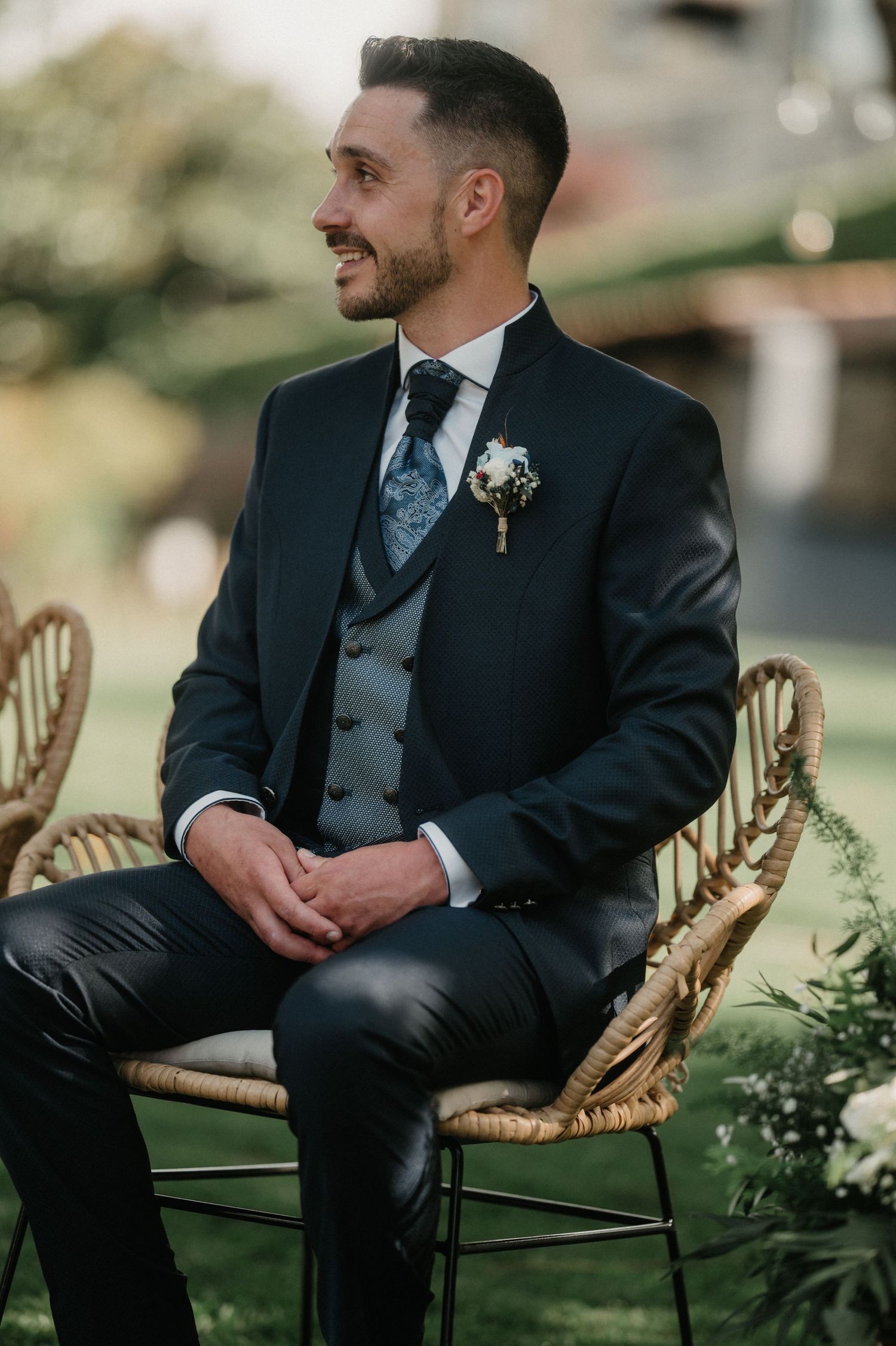 Groom in classic black suit seated on rattan chair during outdoor wedding ceremony.