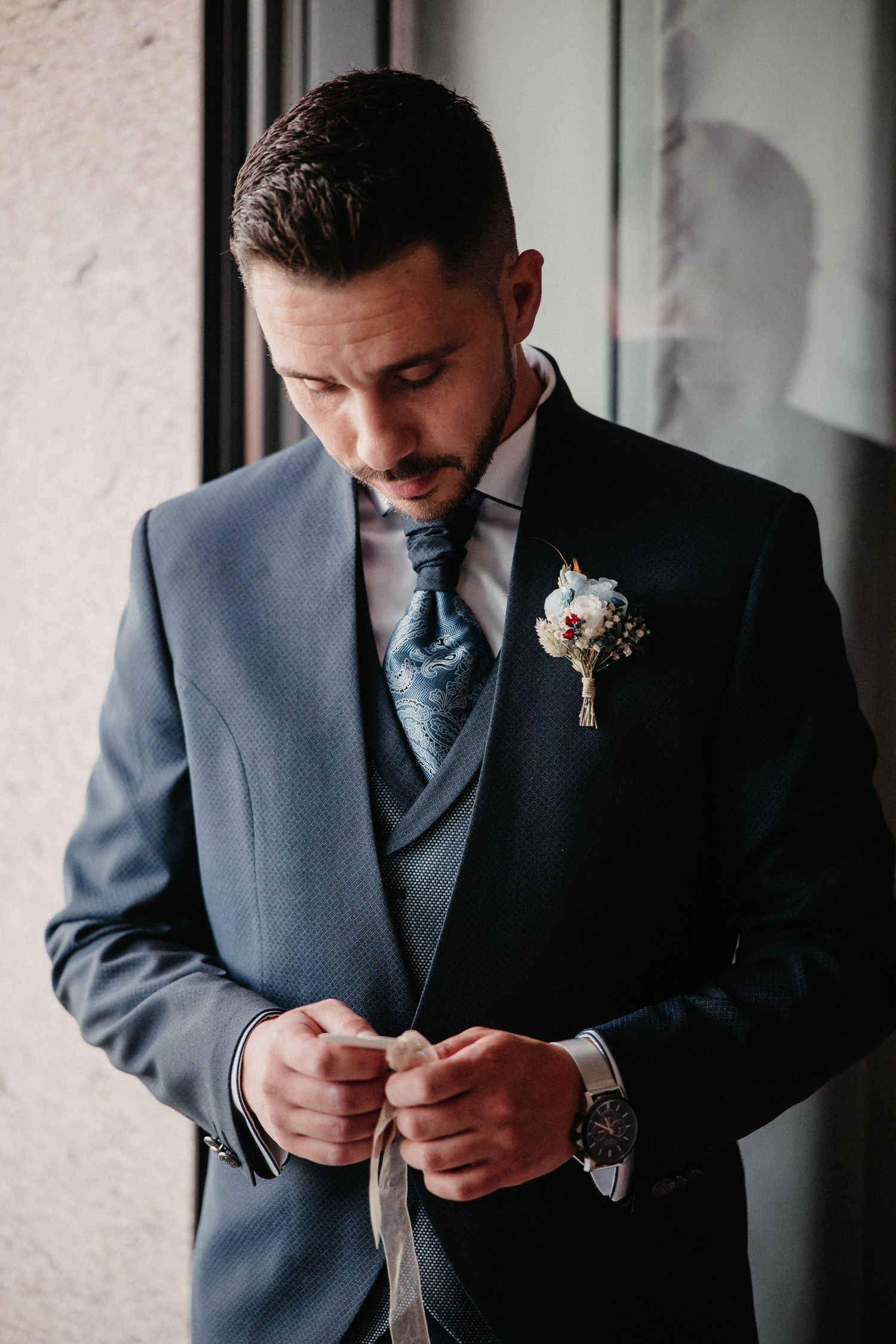 A person in a gray suit and blue tie adjusts their cufflinks while getting ready for a formal event.