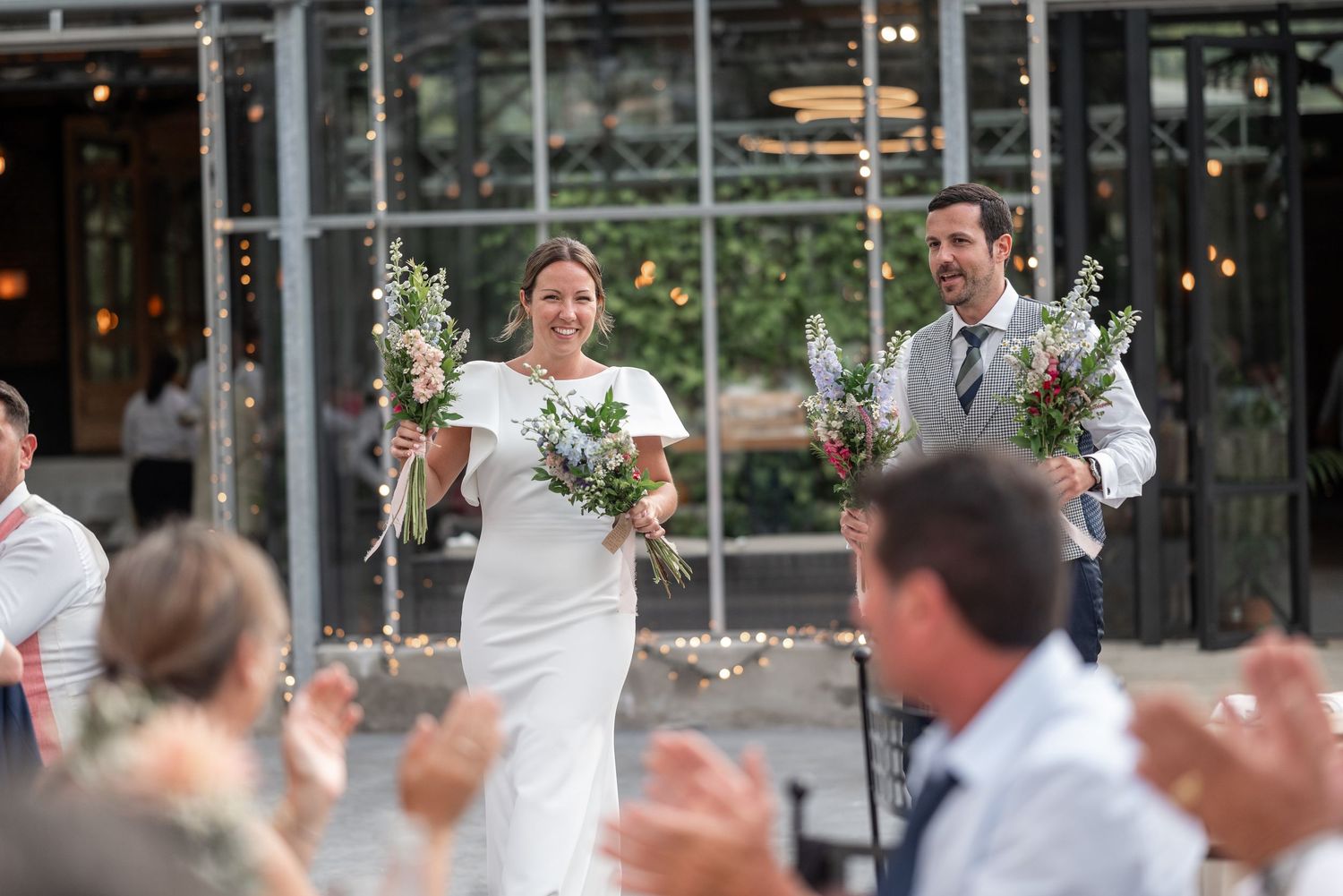 Wedding guests celebrate and applaud during an outdoor reception with string lights overhead.
