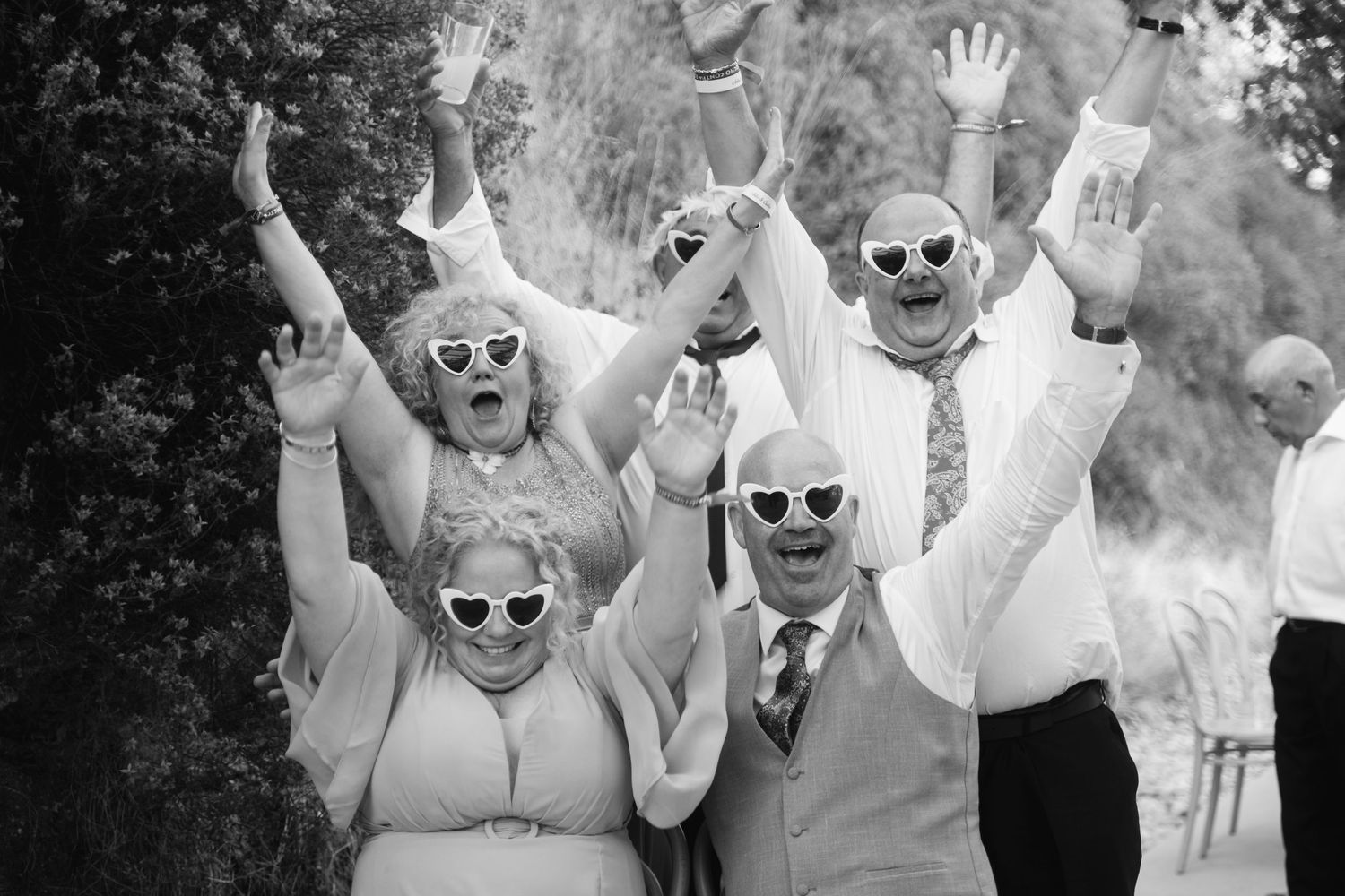 Wedding party wearing matching sunglasses raise their hands joyfully in a black and white celebration photo.