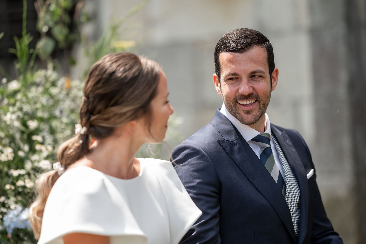 Newlyweds share laughter while sitting together outdoors surrounded by greenery and stone architecture.
