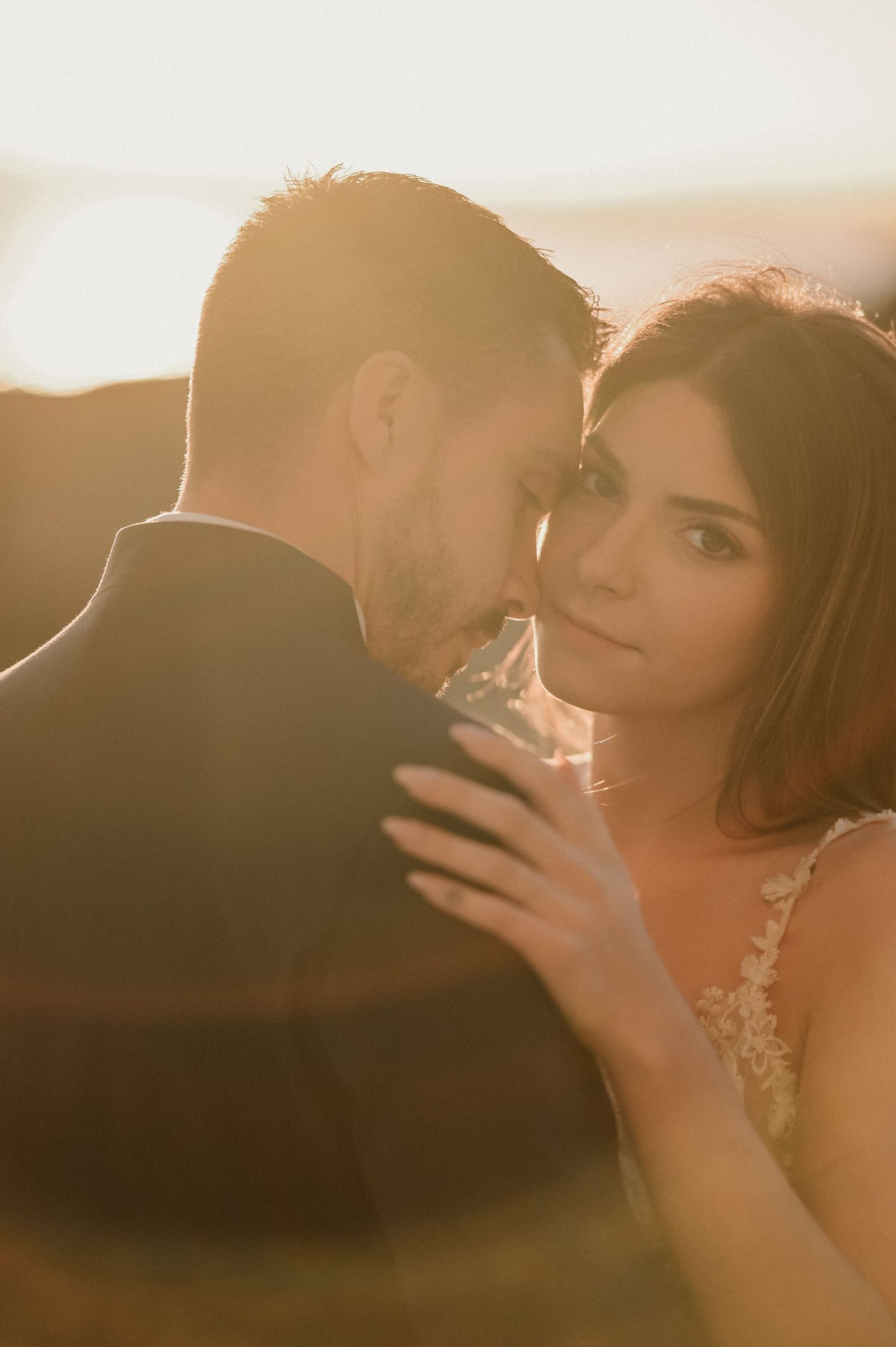 Romantic couple embraces in warm sunset light with bride wearing lace dress during intimate wedding photoshoot.