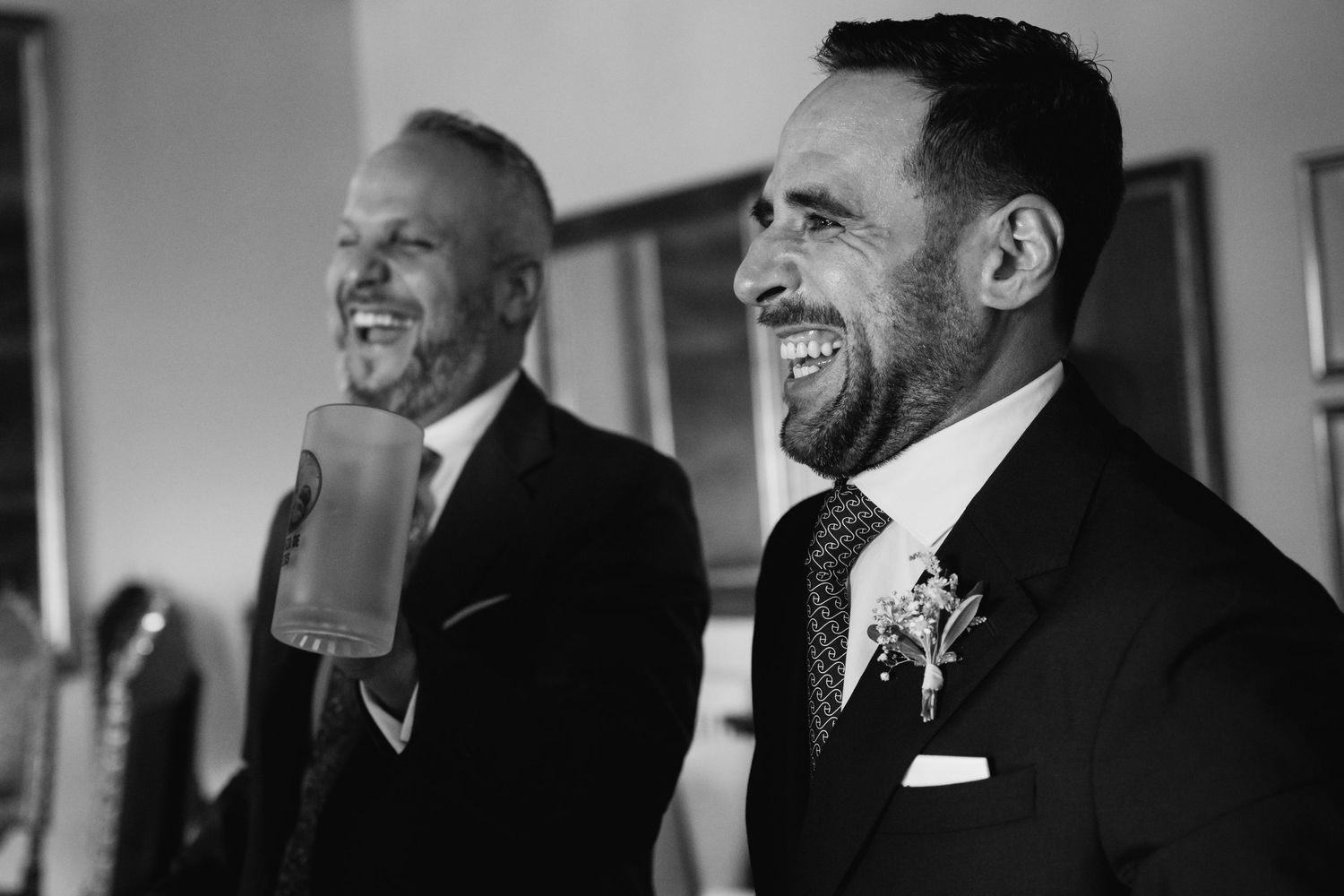 Black and white candid photo of two men in suits laughing together during a wedding celebration.