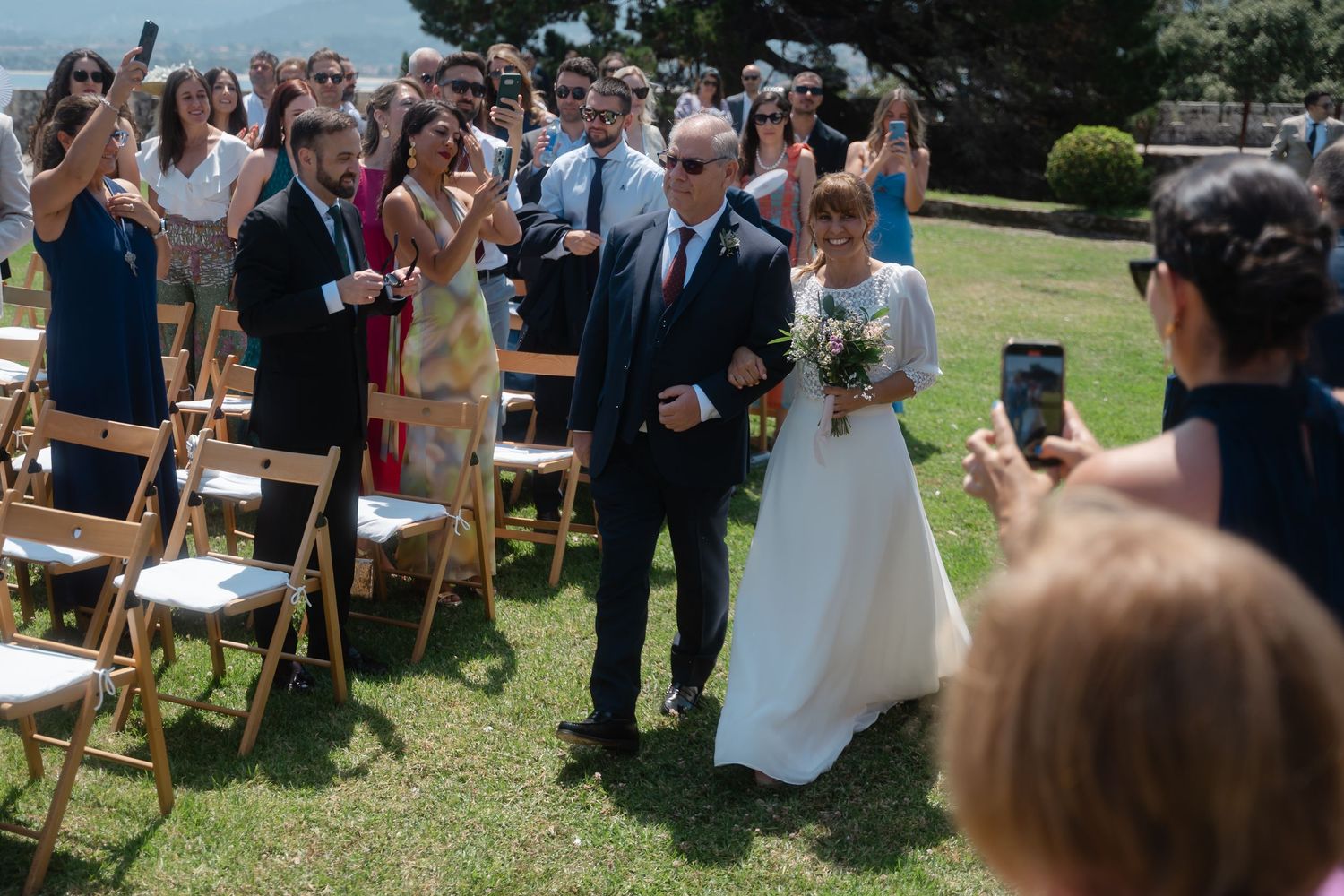 Wedding guests stand on a grassy lawn during an outdoor ceremony on a sunny summer day.
