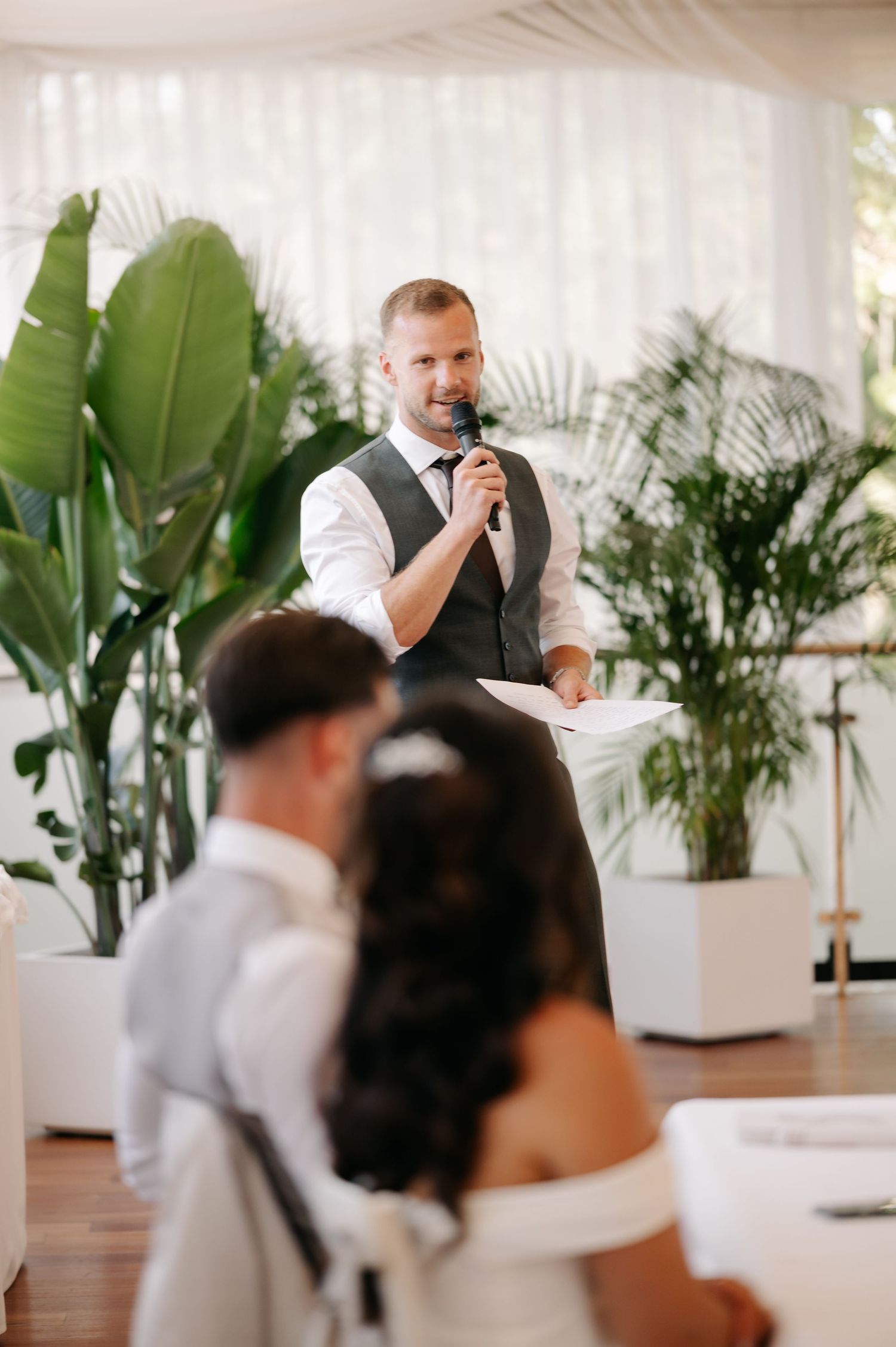 A speaker addresses guests at an elegant indoor venue decorated with tropical plants and white decor.