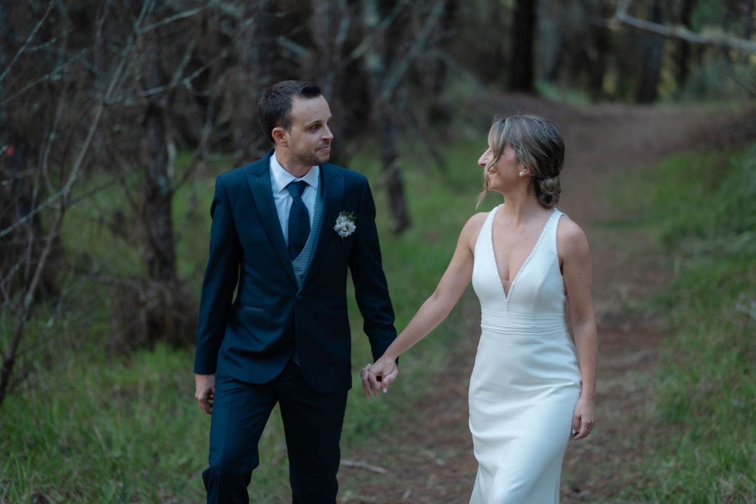 A couple in wedding attire walks together through a wooded outdoor setting.