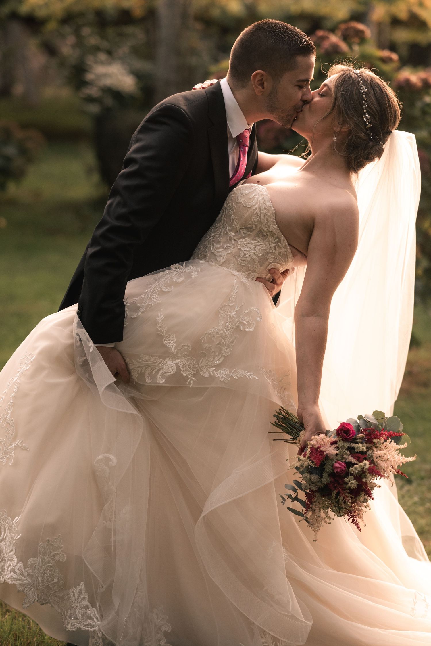 A romantic wedding couple shares a kiss while the bride holds a pink bouquet in an outdoor garden setting.