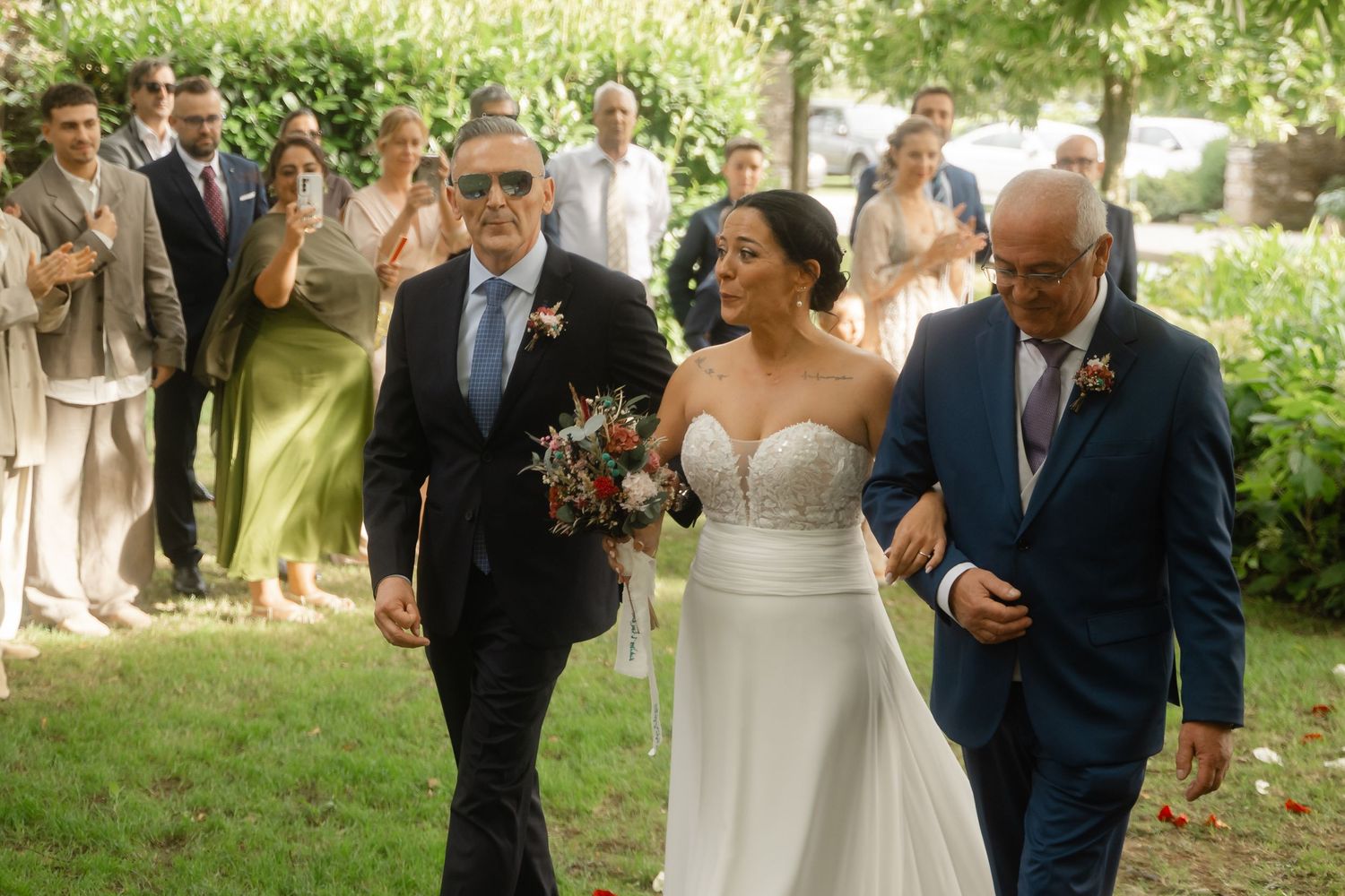 A couple walks together outdoors during a wedding ceremony while guests look on and take photos in a garden setting.