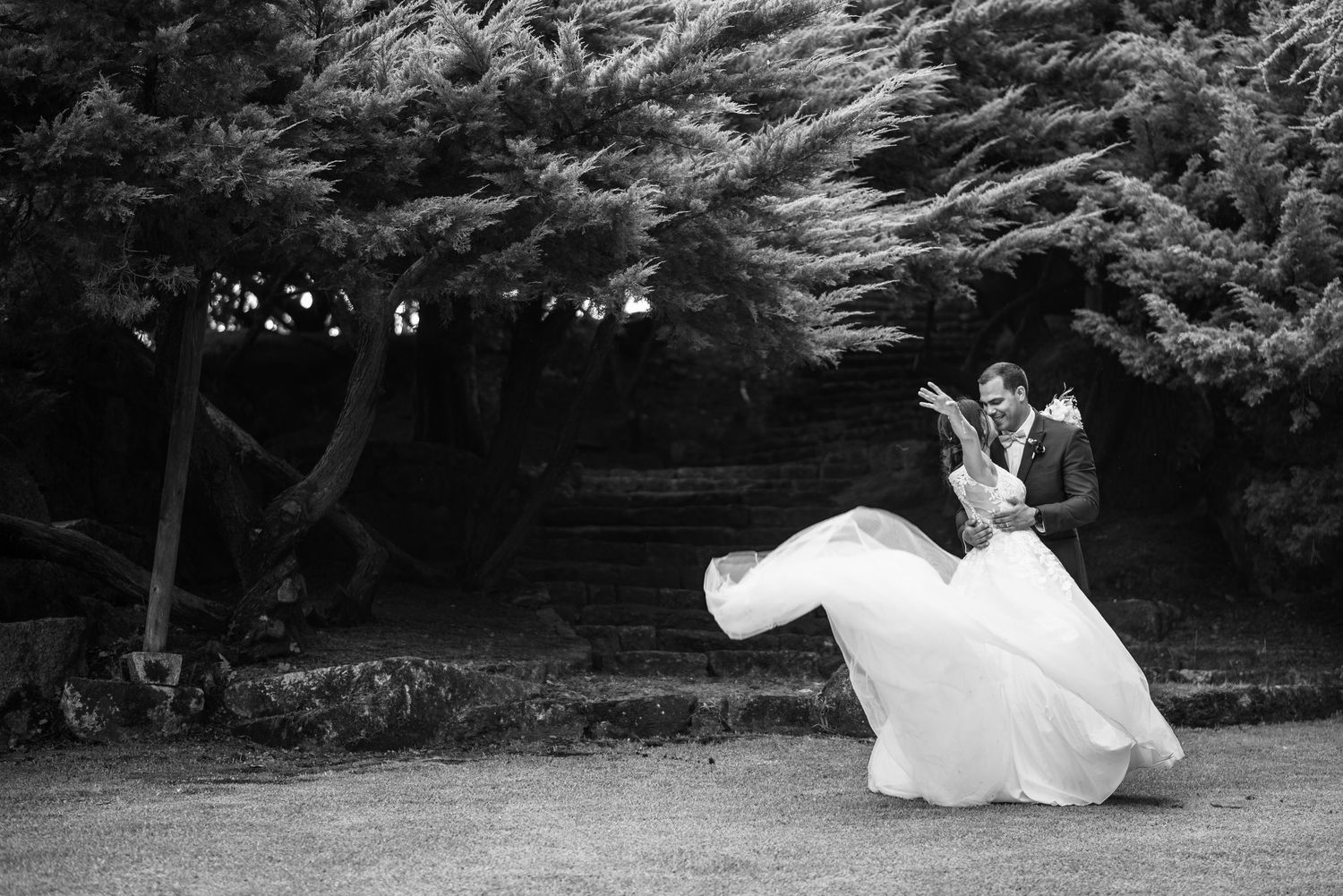 A romantic black and white photograph of a couple dancing outdoors beneath trees in a garden setting.