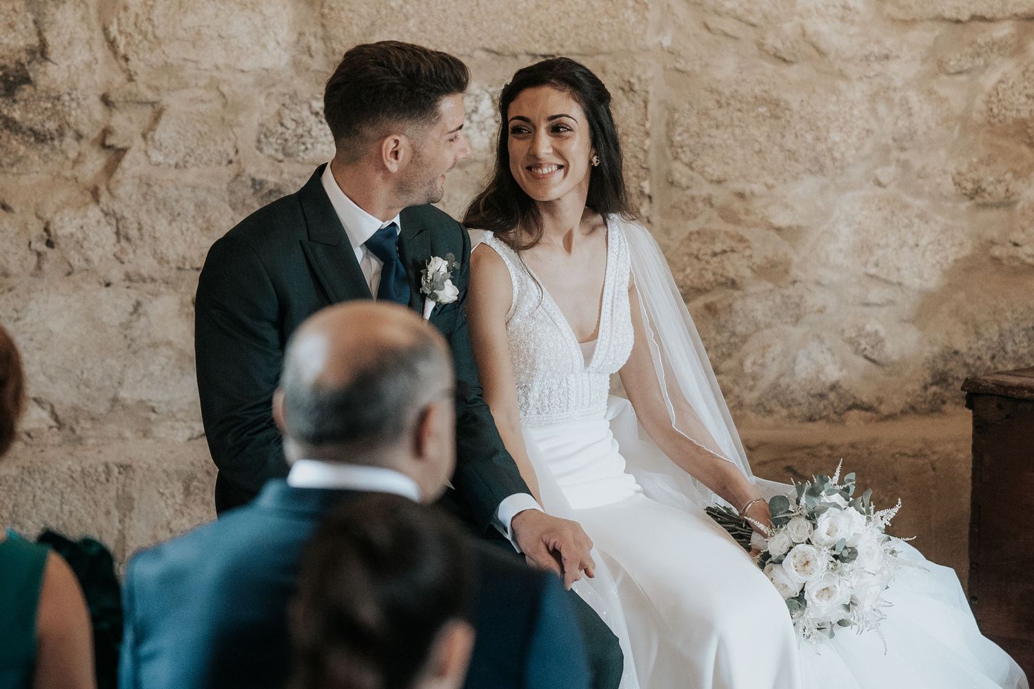 Romantic wedding couple shares intimate moments in rustic stone venue with soft natural lighting and white floral bouquet.