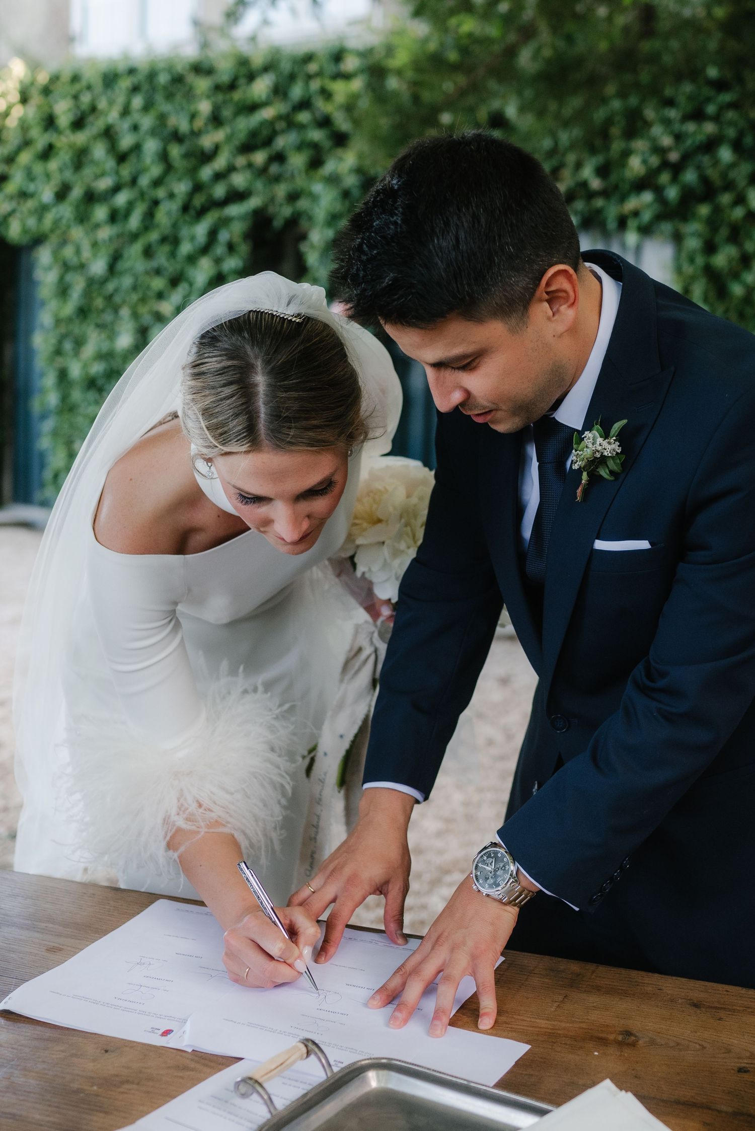 A bride and groom sign their marriage license together at a wooden table during their wedding ceremony.