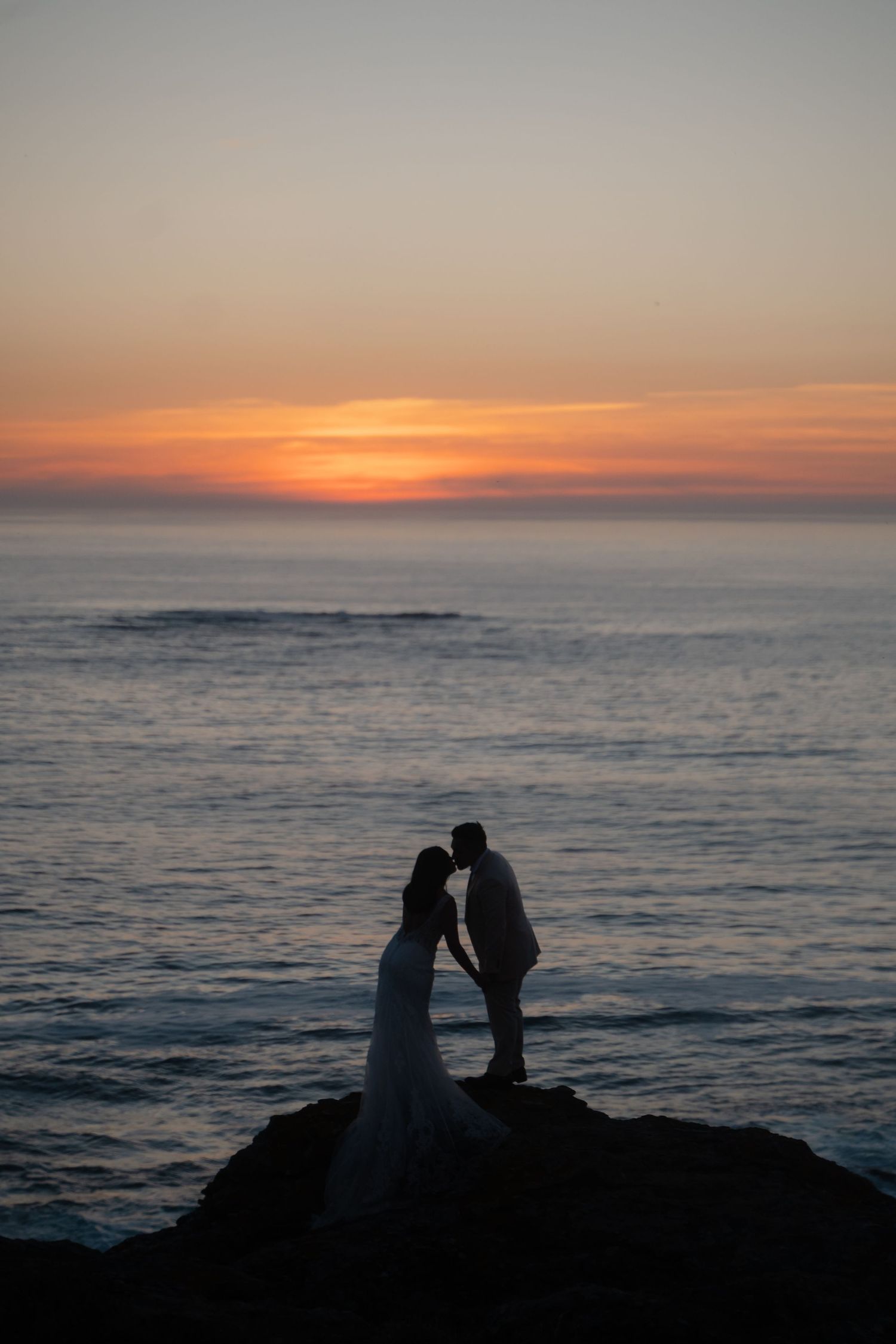 A silhouetted couple shares an intimate moment on coastal rocks during a vibrant sunset.