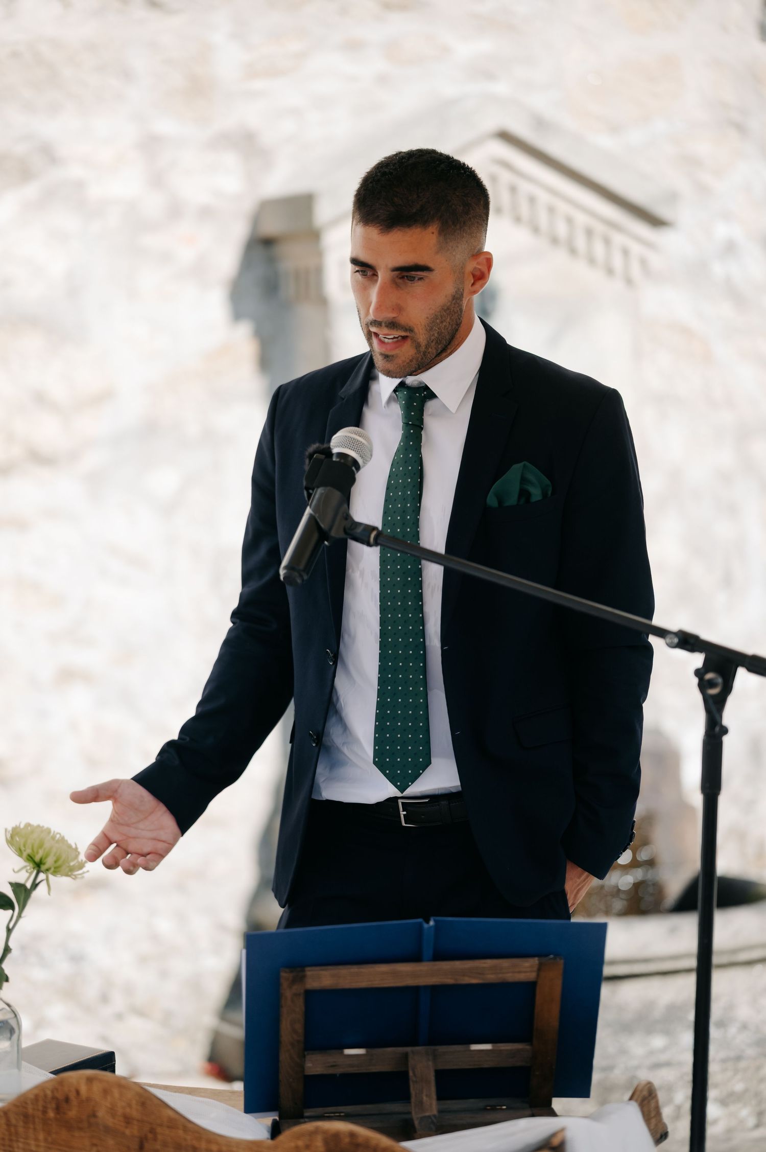 A speaker in a dark suit and green tie sits at a wooden podium during a formal event.