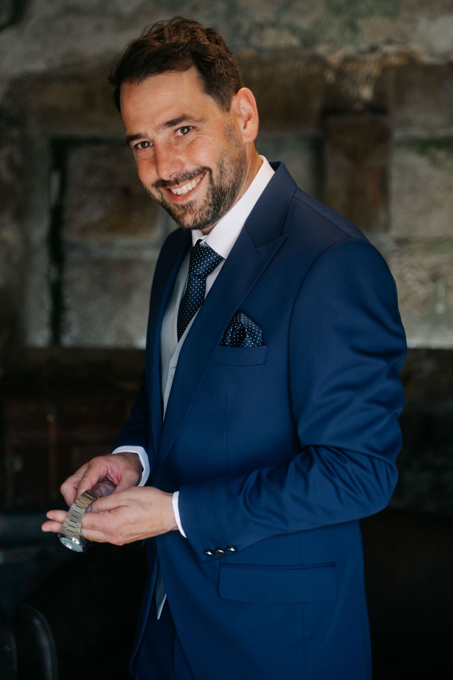 A person wearing a navy blue suit adjusts their cufflinks while standing in a dark, textured setting.