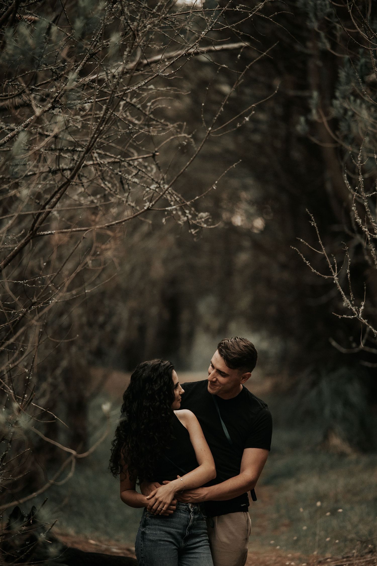 Couple sharing an intimate moment among bare tree branches in a moody forest setting.