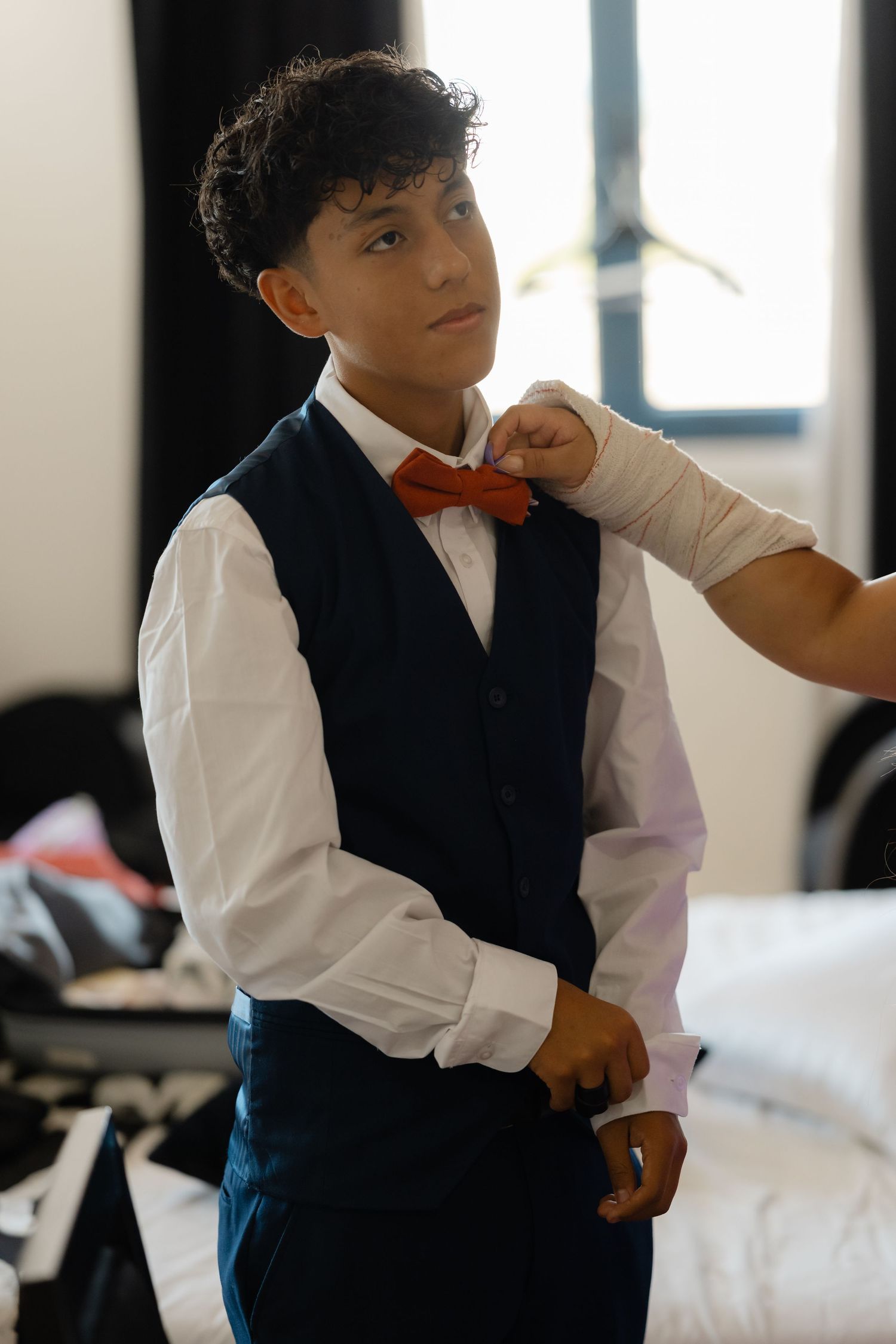 Someone getting ready for a formal event wearing a white dress shirt, navy vest and red bow tie while being helped with attire.