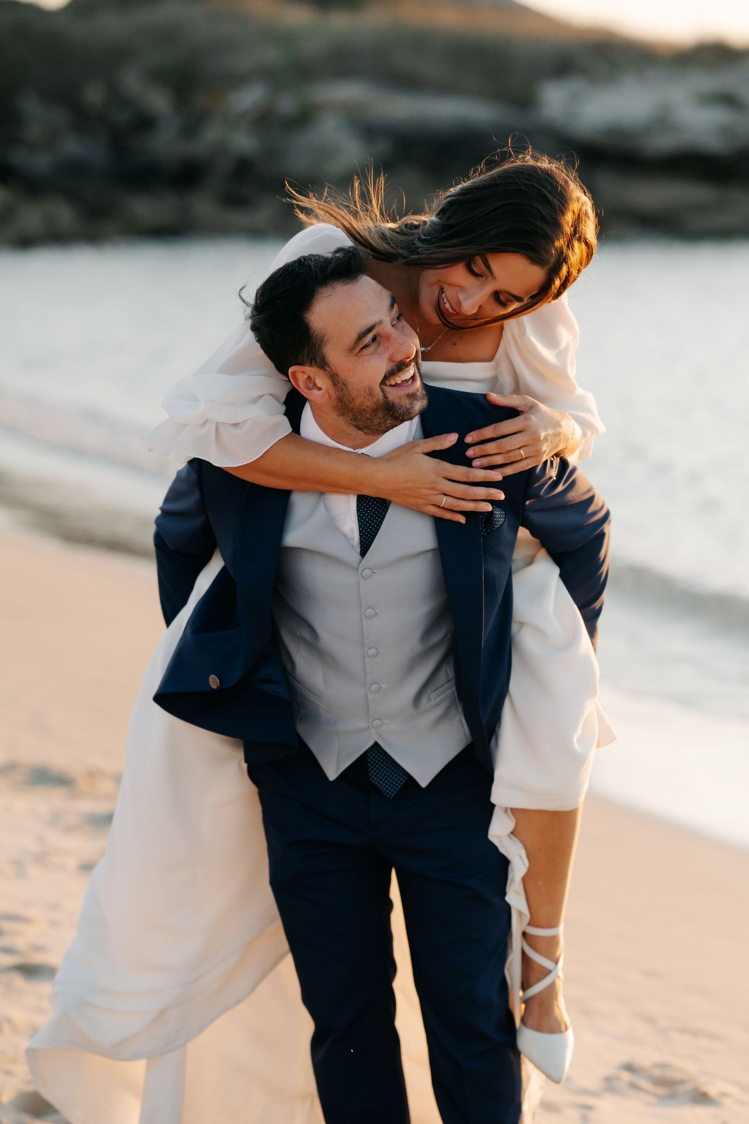 A couple shares a playful moment on the beach at sunset, with the groom in a navy suit and grey vest.