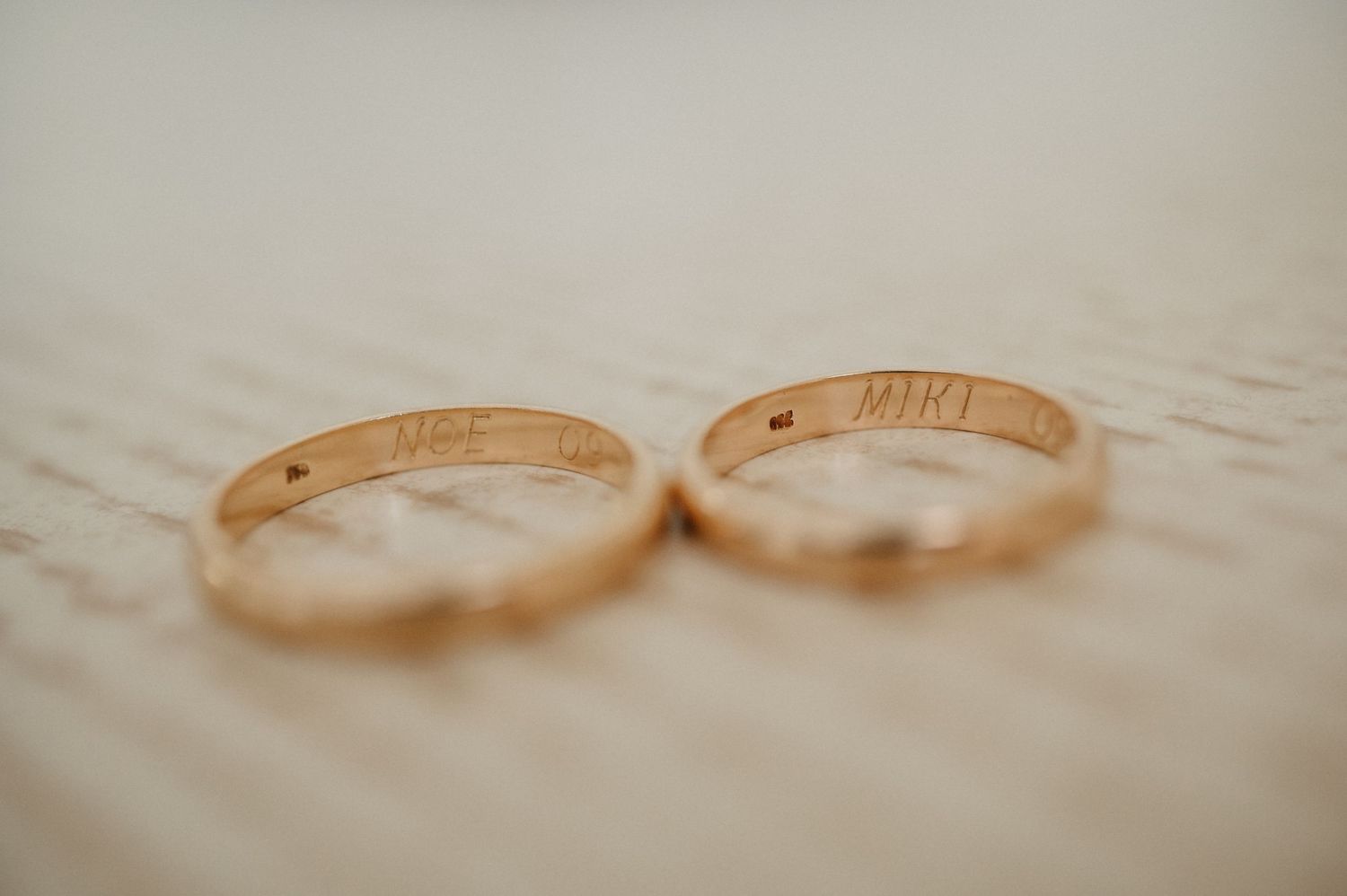 Two gold wedding bands with engraved inscriptions rest on a textured cream-colored surface.