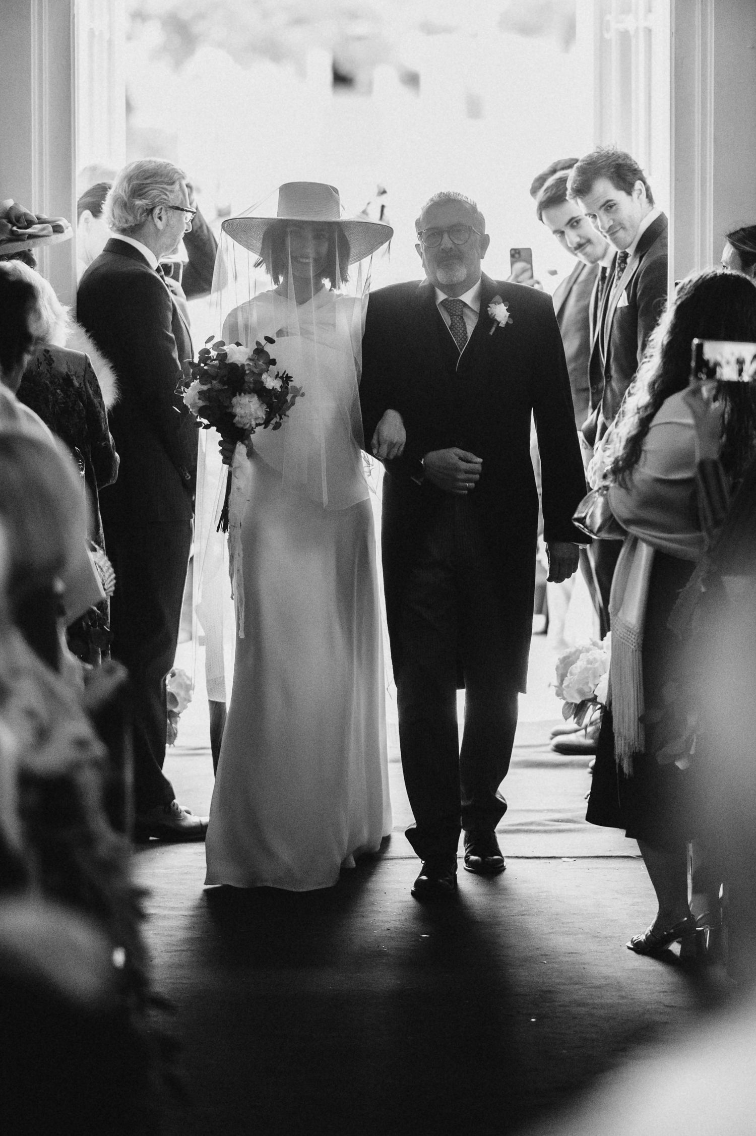 Black and white silhouettes of wedding guests and a bride in a veil walking through a bright interior space.