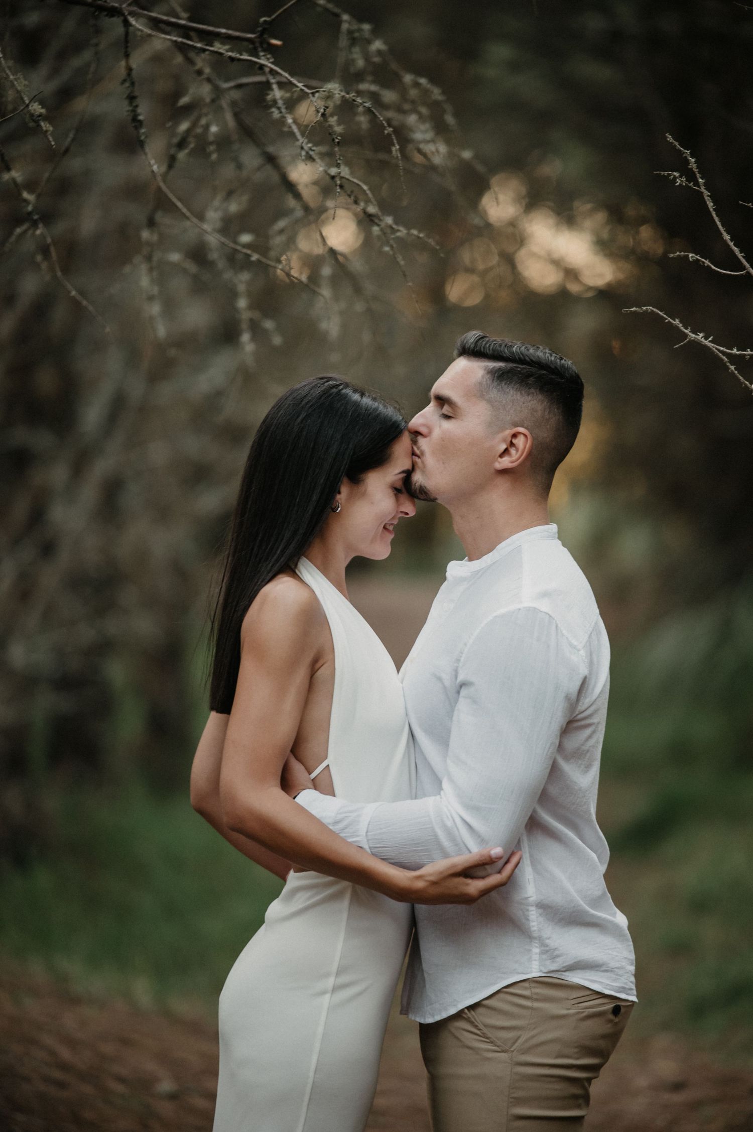 Romantic couple in white embracing in a dreamy forest setting with soft bokeh lighting in the background.