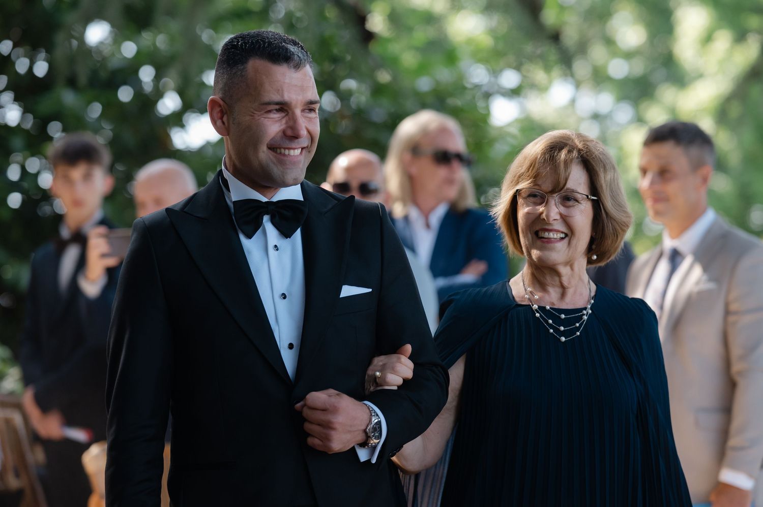 Wedding guests laugh and smile during an outdoor celebration in formal attire.