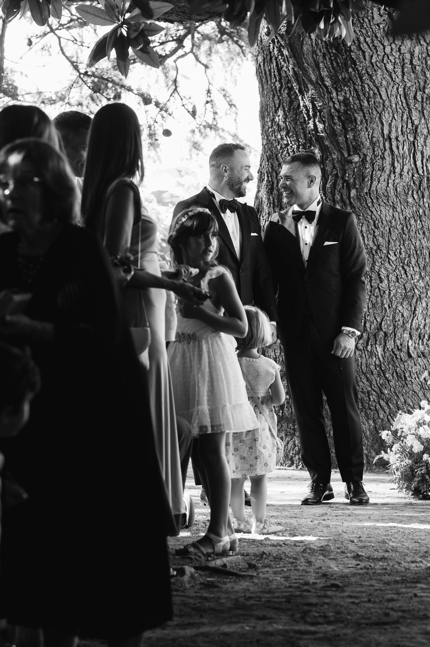 Black and white wedding guests gather under large trees during an outdoor ceremony, creating a candid silhouette effect.