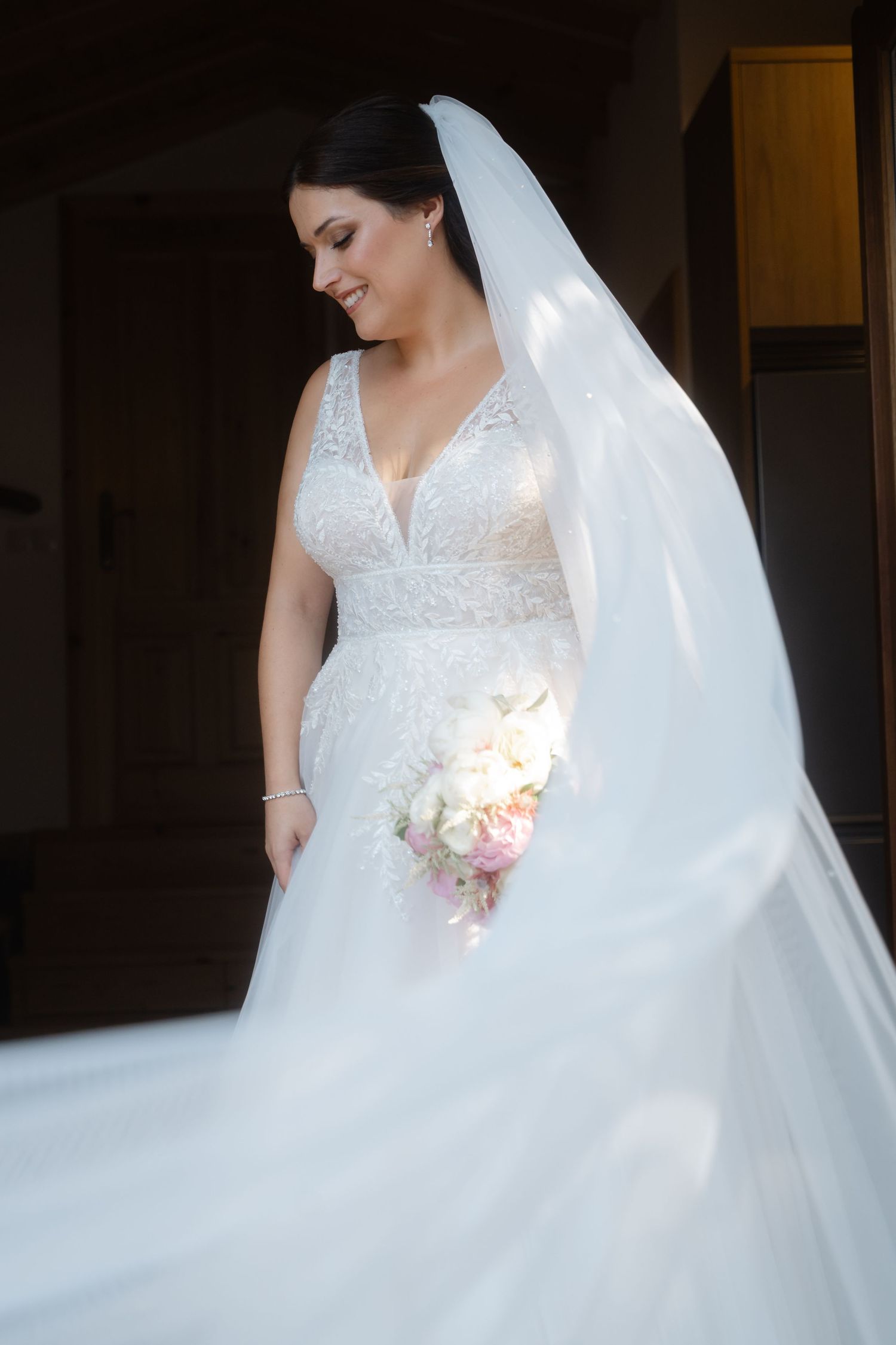 Elegant white wedding gown with flowing veil and delicate bouquet captured in soft indoor lighting.
