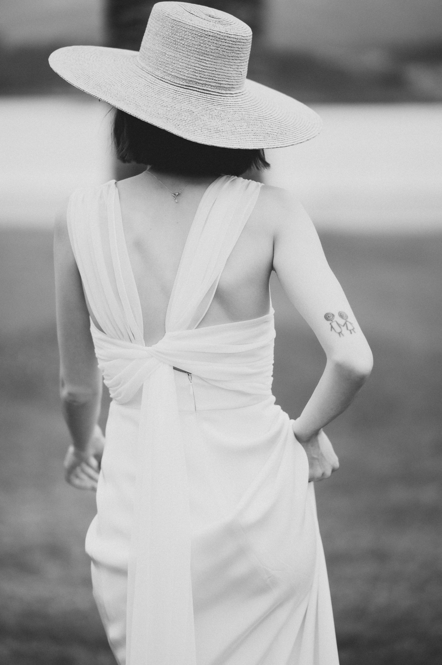 A bride in a white dress and wide-brimmed hat is captured in black and white from behind.