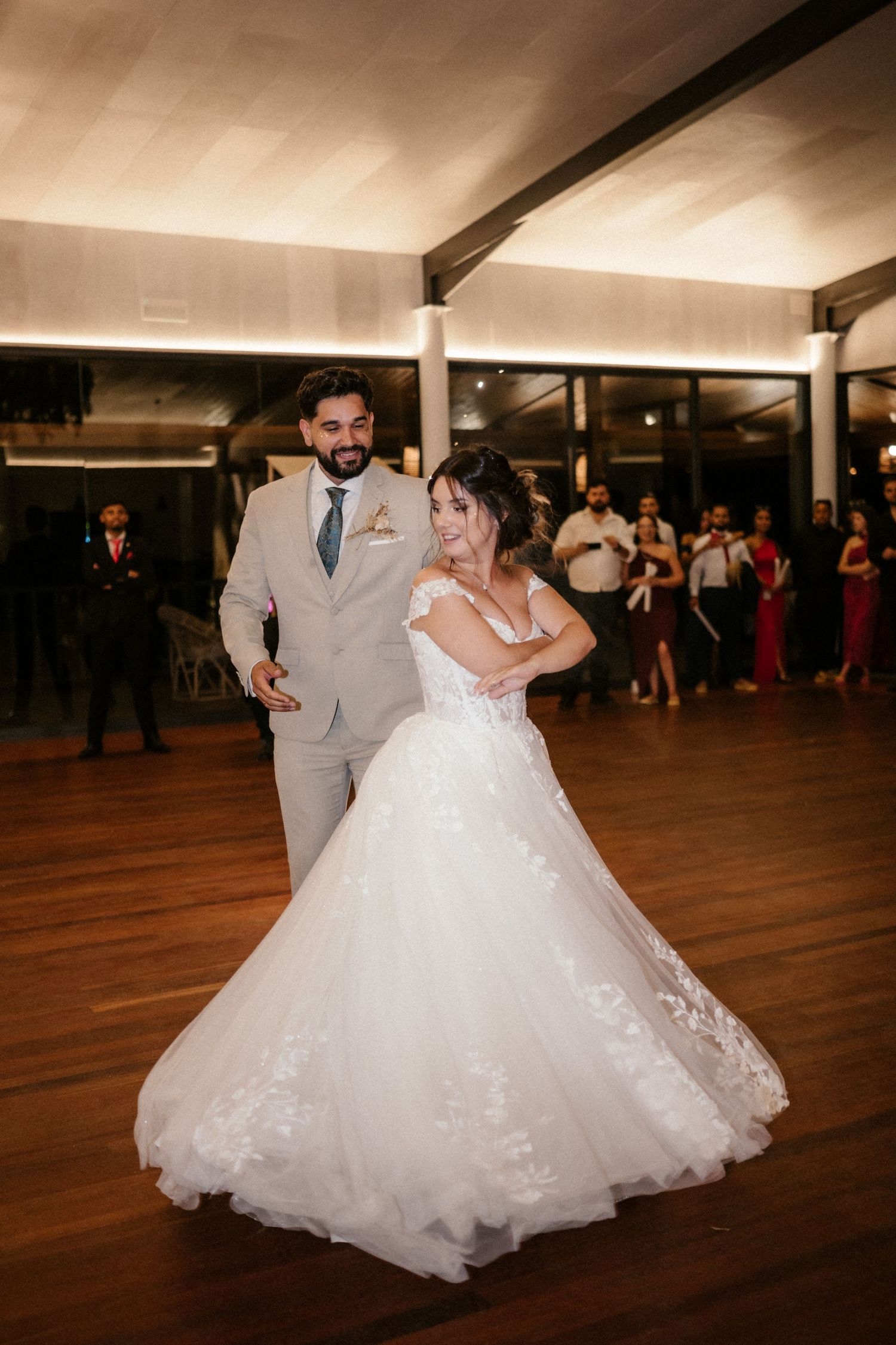 A couple shares their first dance at their wedding reception on a wooden dance floor with guests watching in the background.