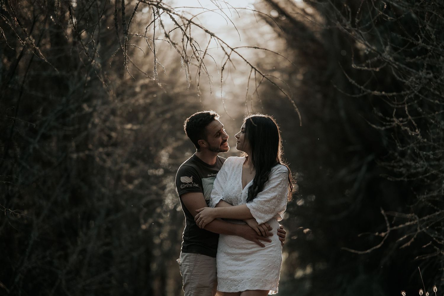 A couple embraces in a moody forest setting with dramatic backlit sunlight filtering through bare branches.