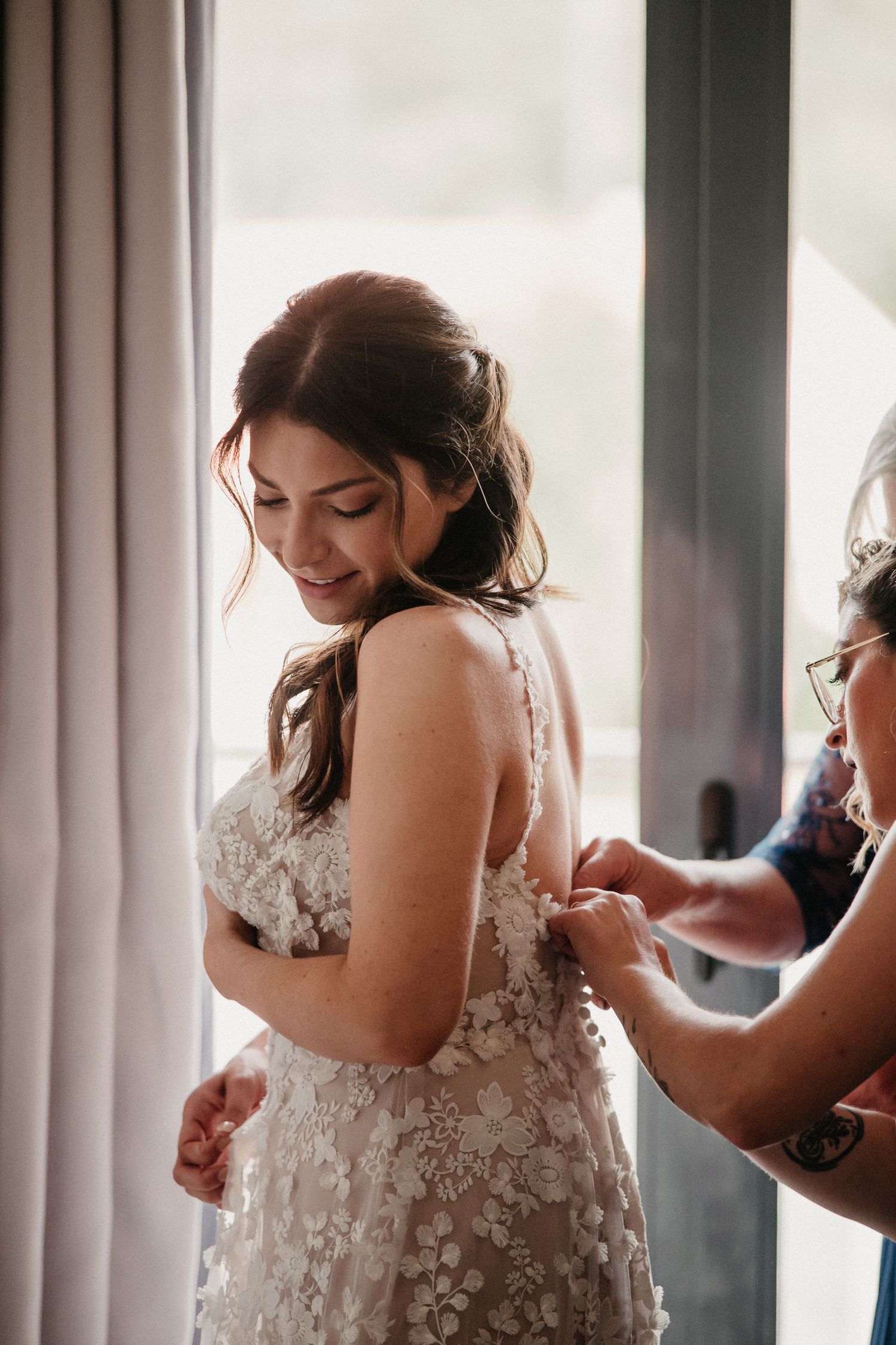Delicate lace wedding dress being fastened in a sunlit room with soft window lighting.