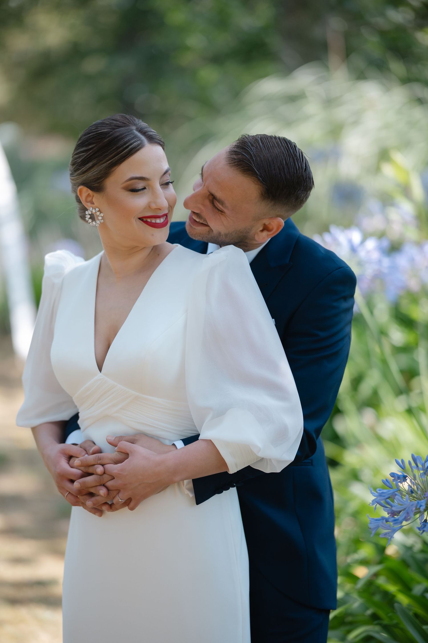 Newlyweds share a tender moment in a garden with blooming purple flowers, bride in elegant white dress.
