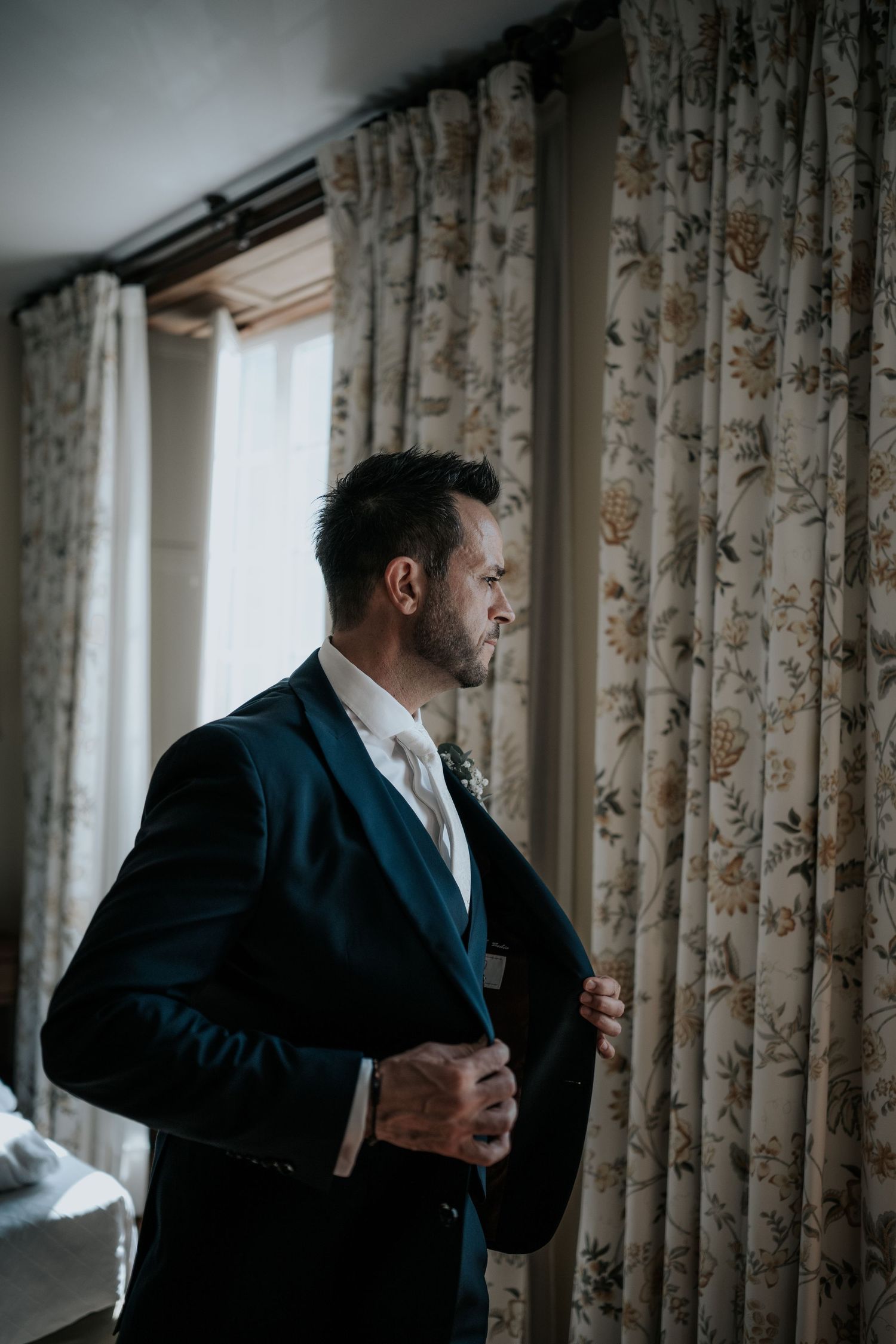 A groom in a dark suit adjusts his tie while standing near floral curtains in natural window light.